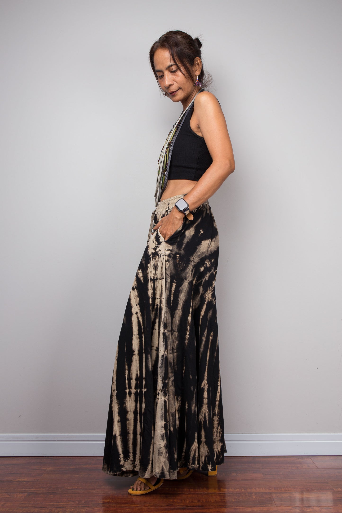 Side view of a model wearing the Nuichan black and sand reverse tie dye wide leg palazzo pants, displaying the full width of the ultra-wide leg silhouette, smocked elastic waistband, the inseam pockets, and the continuation of the vertical bleached stripe pattern across both legs