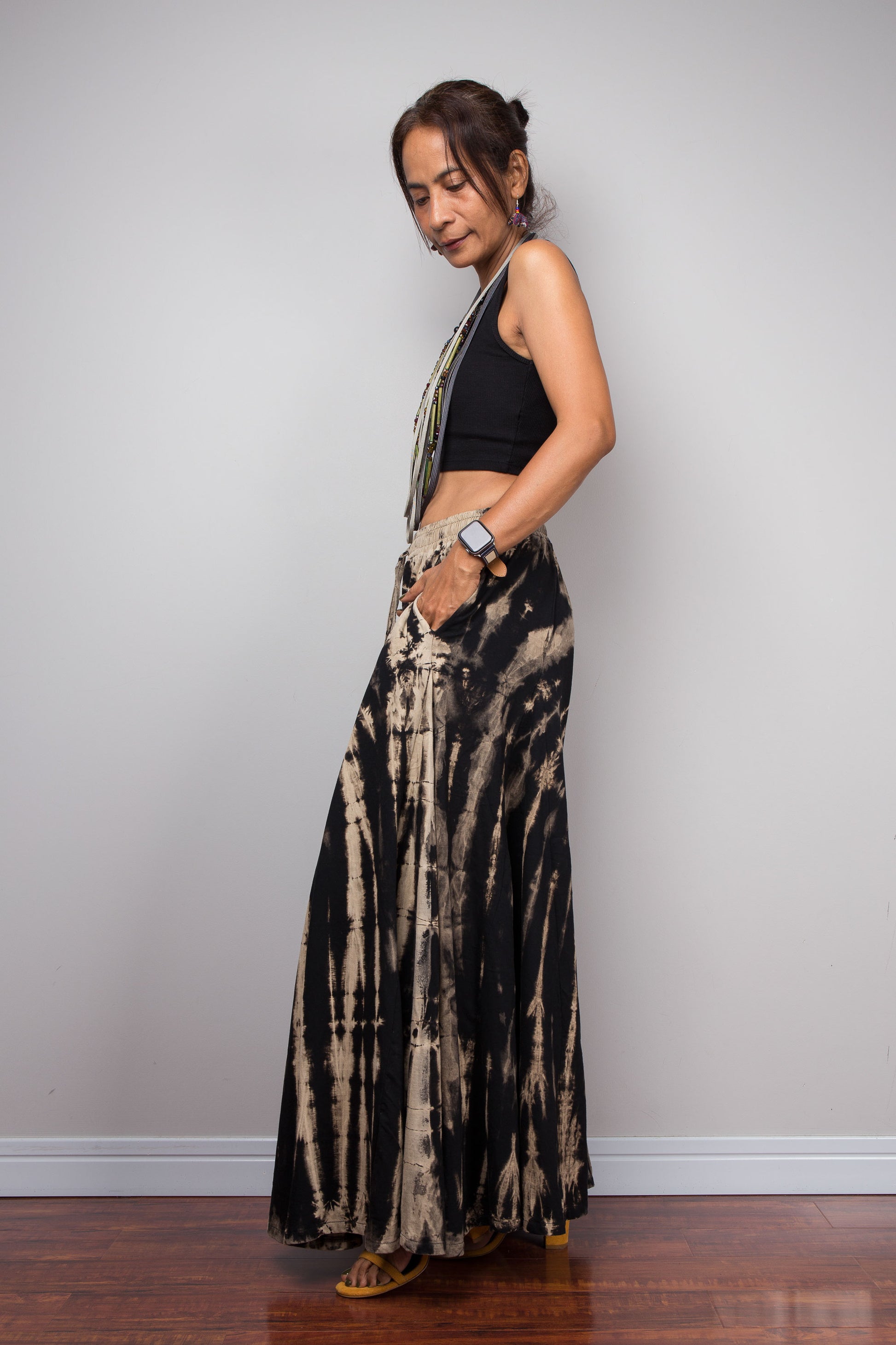 Side view of a model wearing the Nuichan black and sand reverse tie dye wide leg palazzo pants, displaying the full width of the ultra-wide leg silhouette, smocked elastic waistband, the inseam pockets, and the continuation of the vertical bleached stripe pattern across both legs