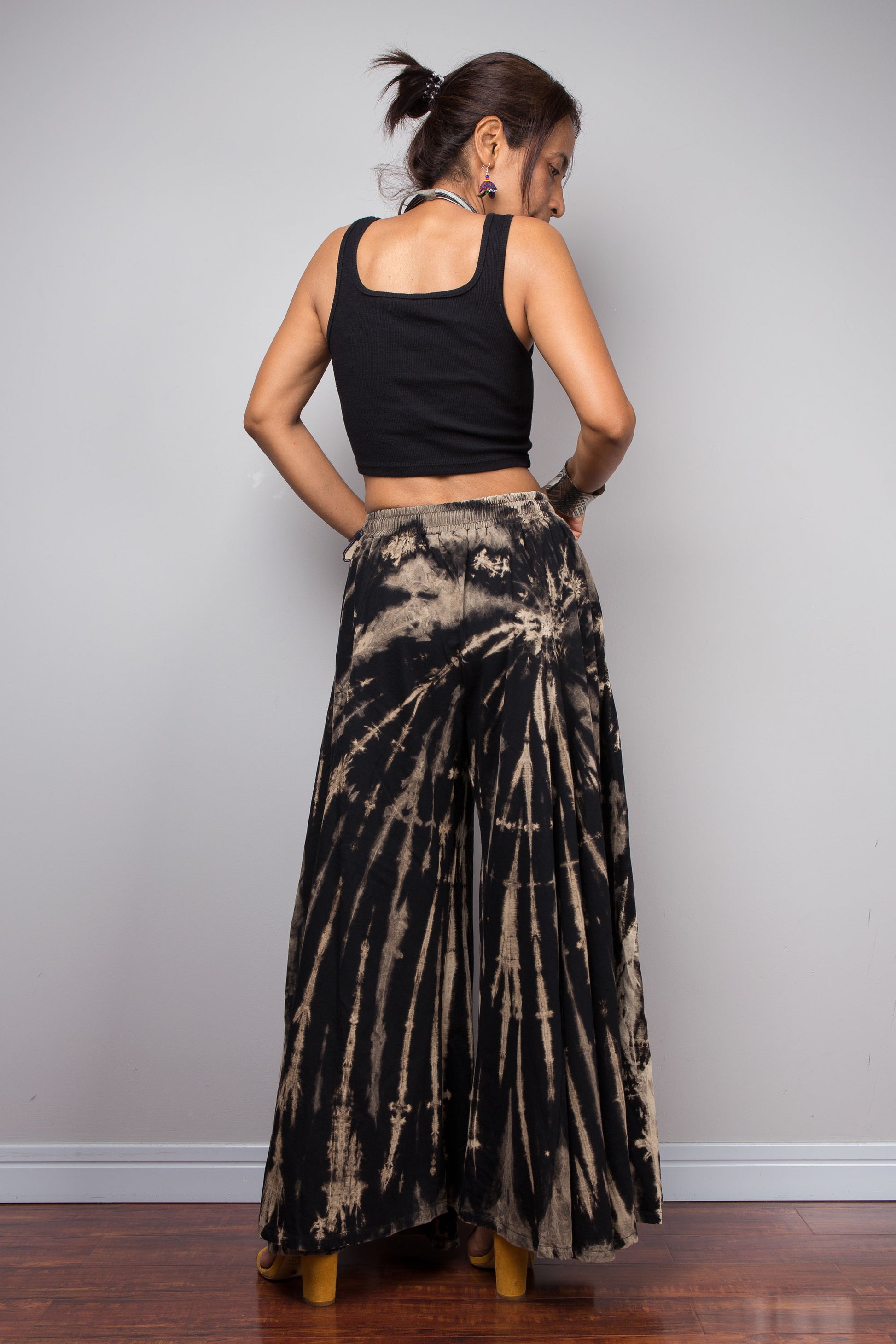 Rear view of a model wearing the Nuichan black and sand reverse tie dye wide leg palazzo pants, displaying the full width of the ultra-wide leg silhouette, smocked elastic waistband with drawstring tie detail at the back, and the continuation of the vertical bleached stripe pattern across both legs
