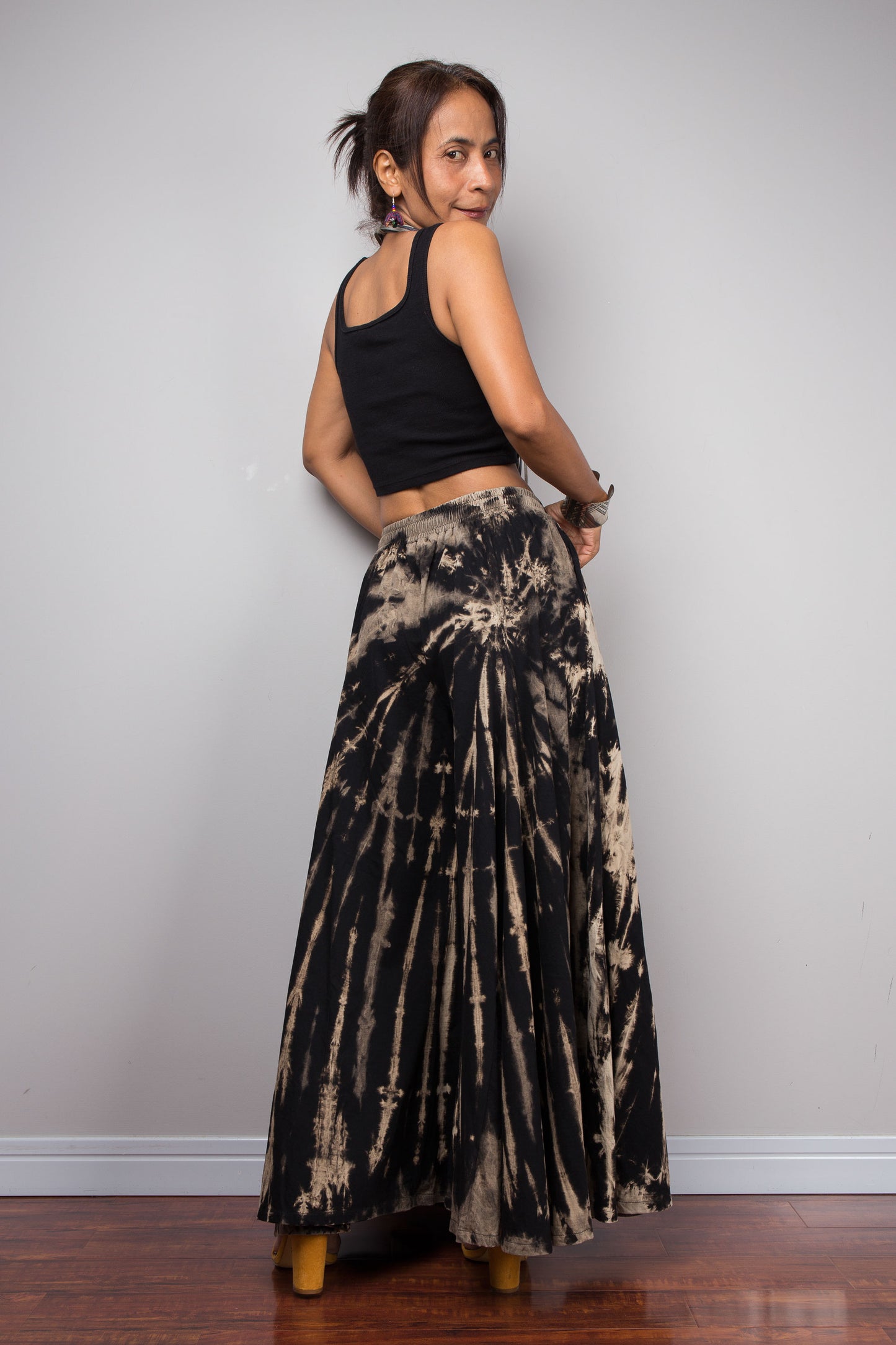 Rear view of a model wearing the Nuichan black and sand reverse tie dye wide leg palazzo pants in a vertical stripe pattern, styled high-waisted with a black crop top, showcasing the dramatic ultra-wide leg silhouette, elongating vertical bleached stripes, and warm sand smocked elastic waistband with adjustable drawstring