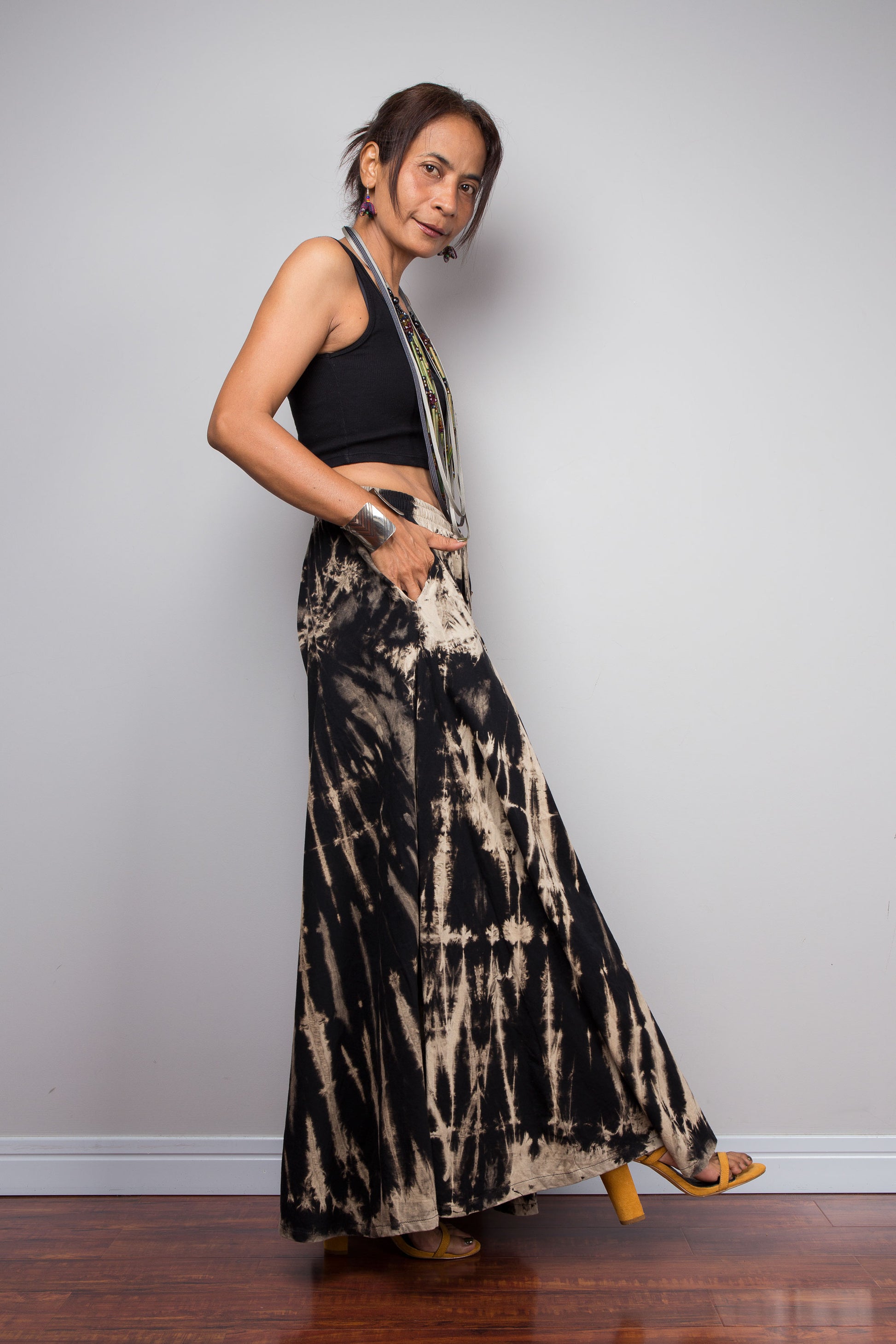 Side view of a petite female model wearing the Nuichan black and sand reverse tie dye wide leg palazzo pants in a vertical stripe pattern, styled high-waisted with a black crop top, showcasing the dramatic ultra-wide leg silhouette, elongating vertical bleached stripes, and warm sand smocked elastic waistband with adjustable drawstring