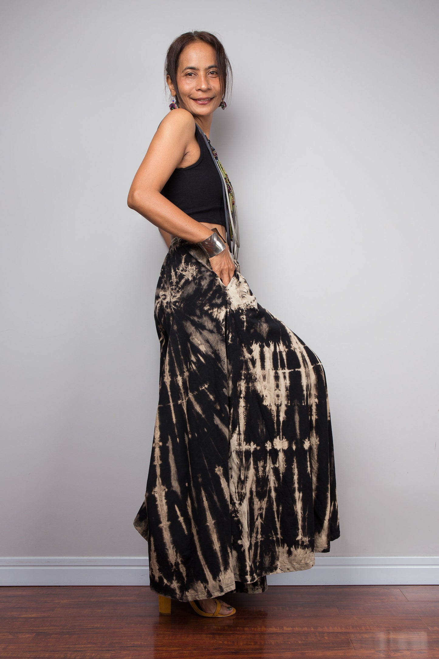 Side view of a model wearing the Nuichan black and sand reverse tie dye wide leg palazzo pants in a vertical stripe pattern, styled high-waisted with a black crop top, showcasing the dramatic ultra-wide leg silhouette, elongating vertical bleached stripes, and warm sand smocked elastic waistband with adjustable drawstring