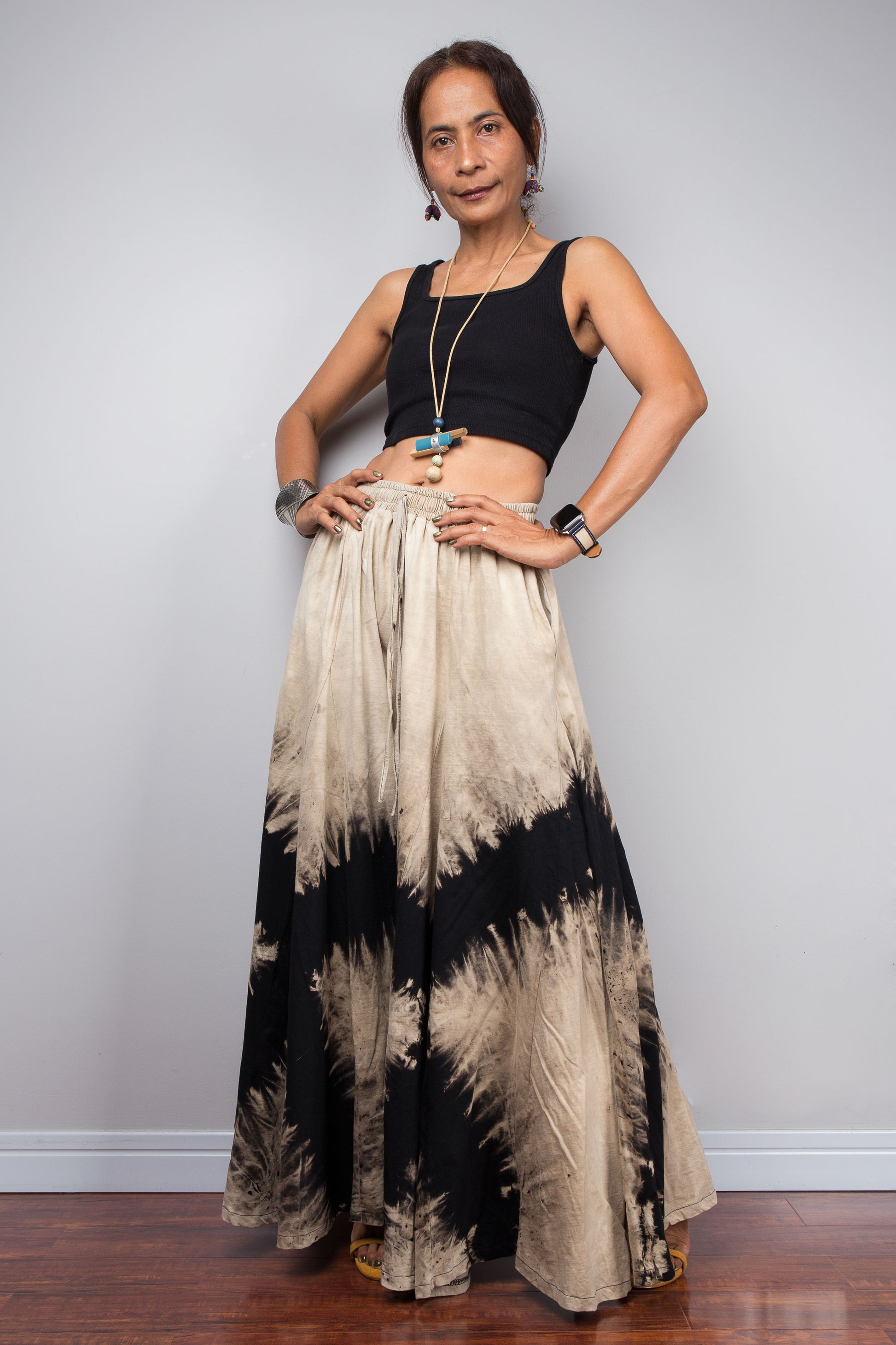 Petite Woman wearing a black and beige reverse tie dye pair of wide leg pants against a white wall.