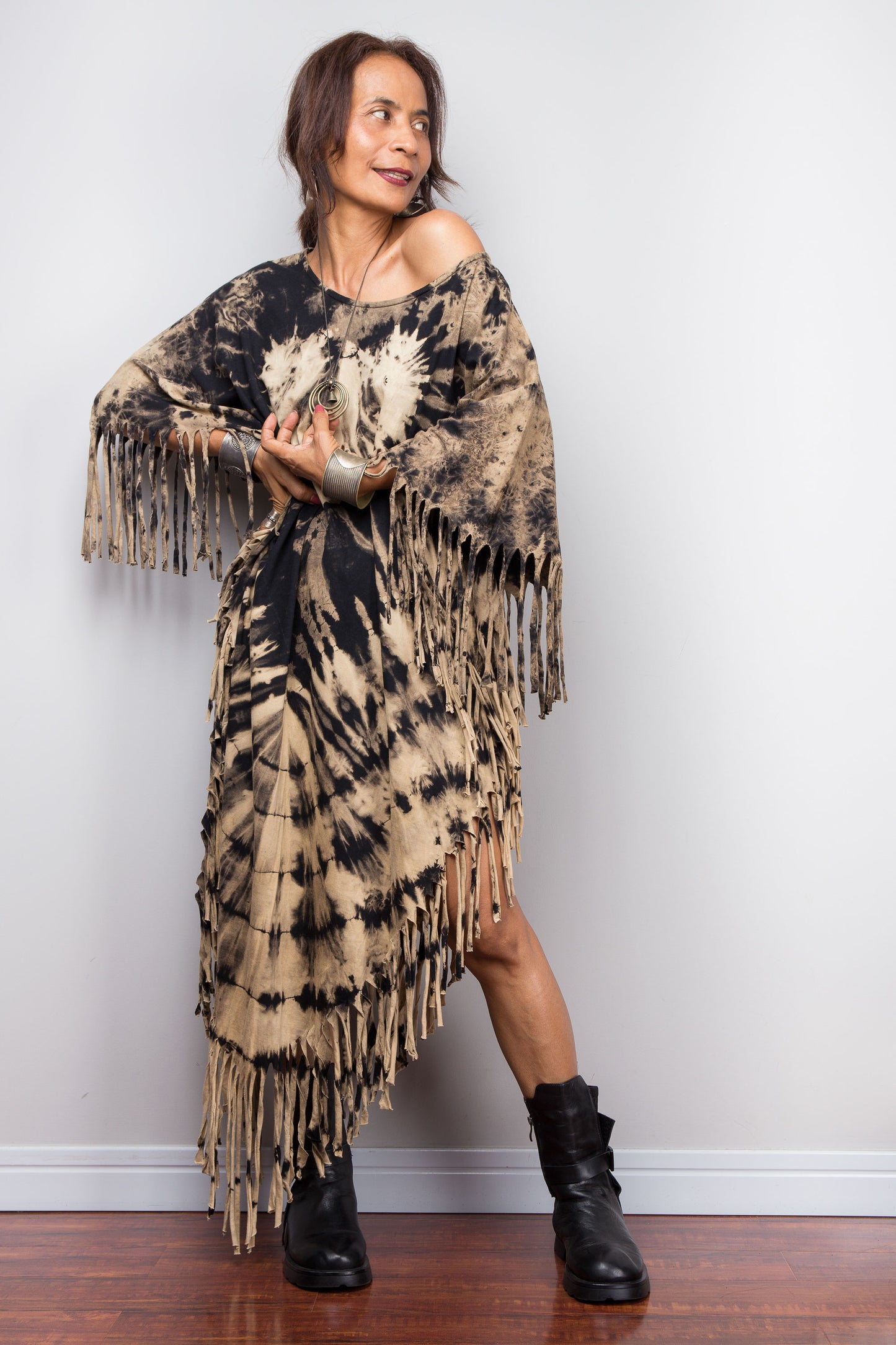 Woman wearing a tie-dye dress with fringes and black boots against a white wall.