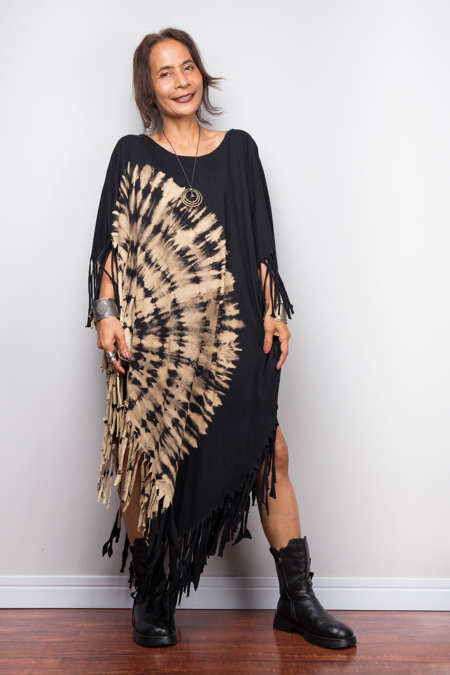 Woman wearing a black and beige tie-dye poncho dress with fringe, standing against a plain white wall.