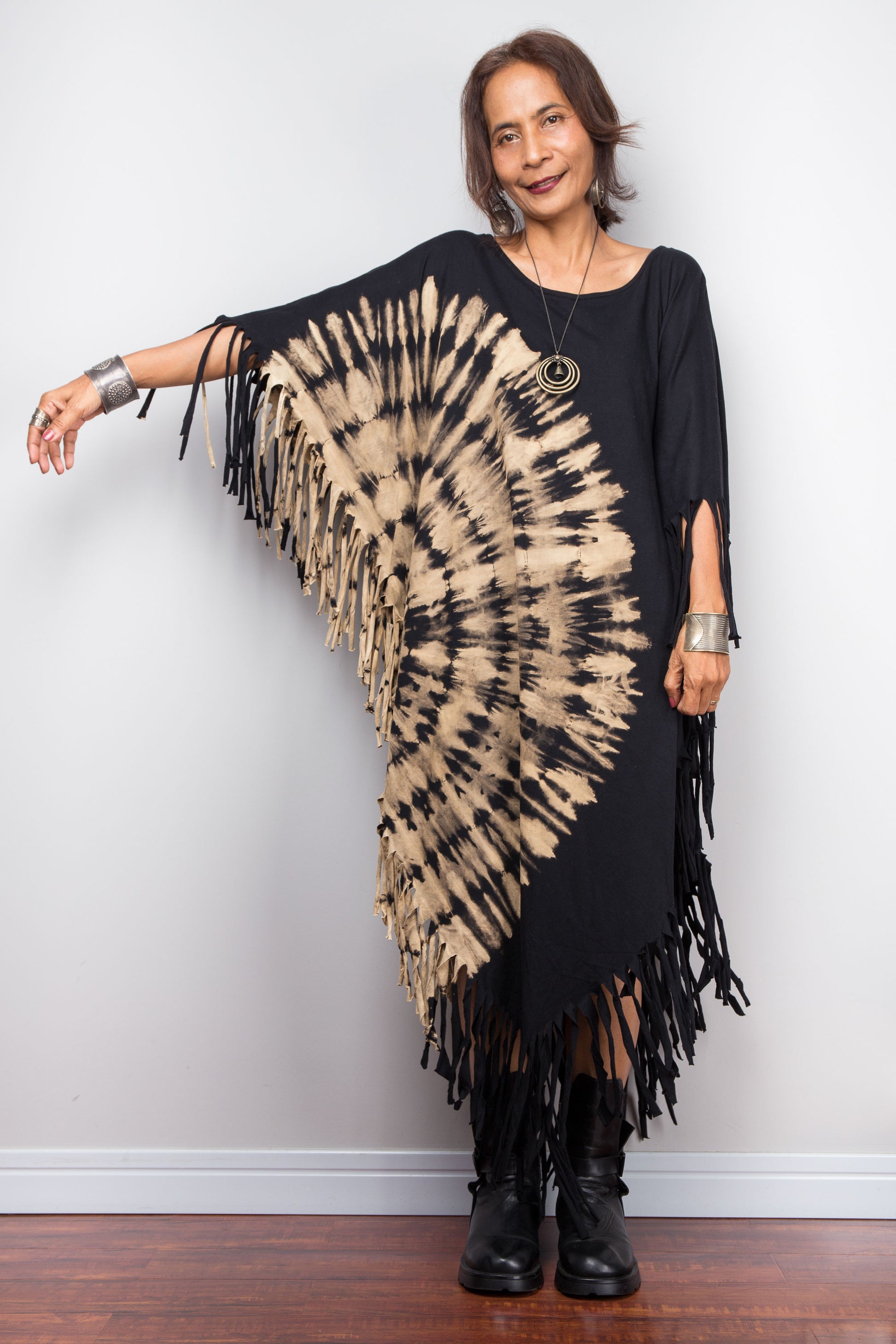 Woman wearing a black and beige tie-dye poncho with fringe standing against a white wall.