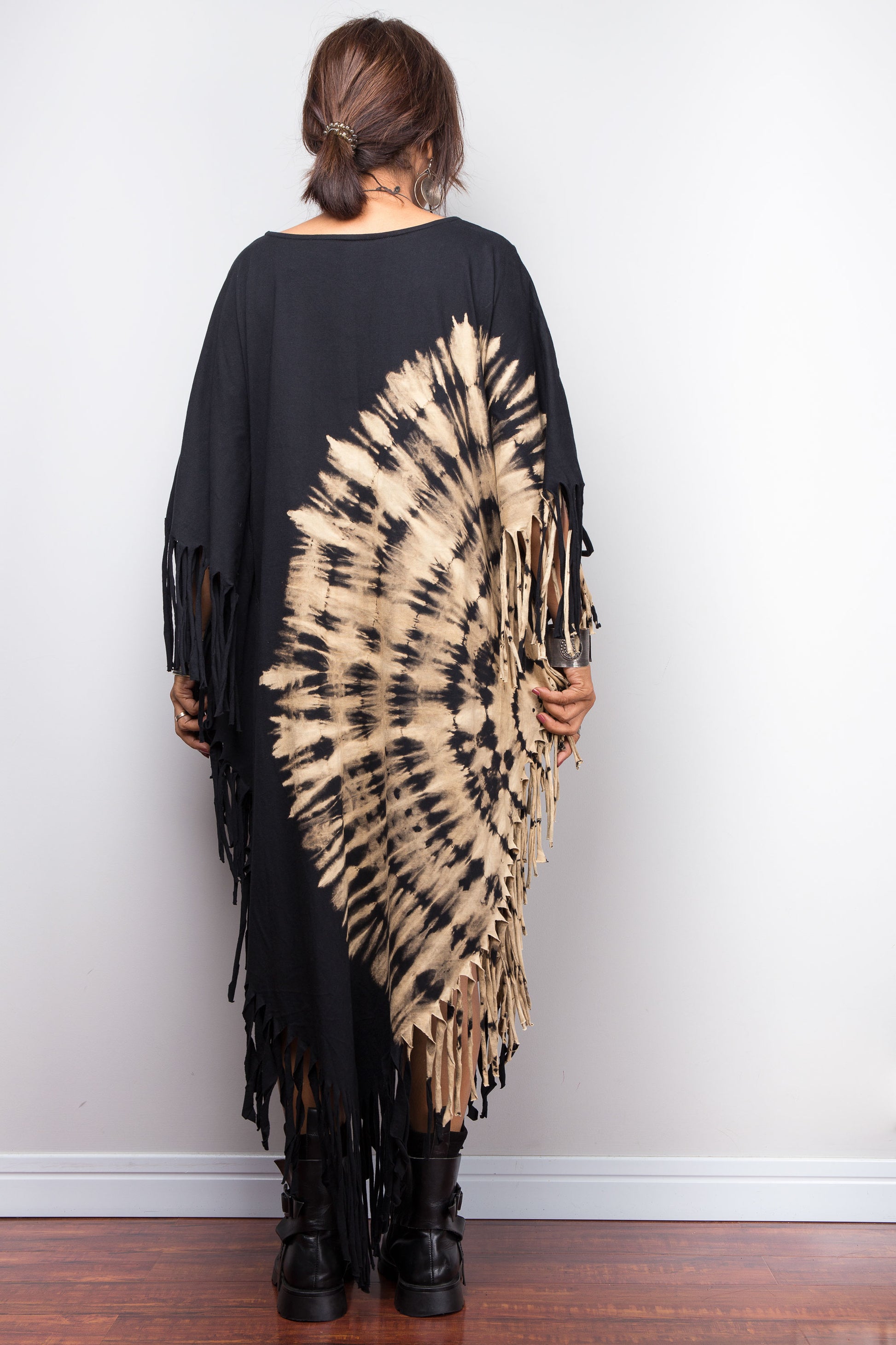 Person wearing a black and beige tie-dye poncho with fringe details on a plain background