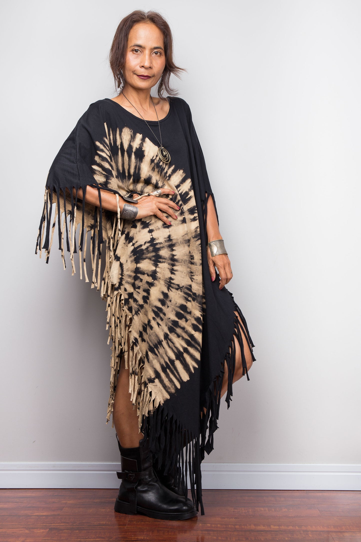 Woman wearing a black and beige tie-dye dress with fringes against a white wall.