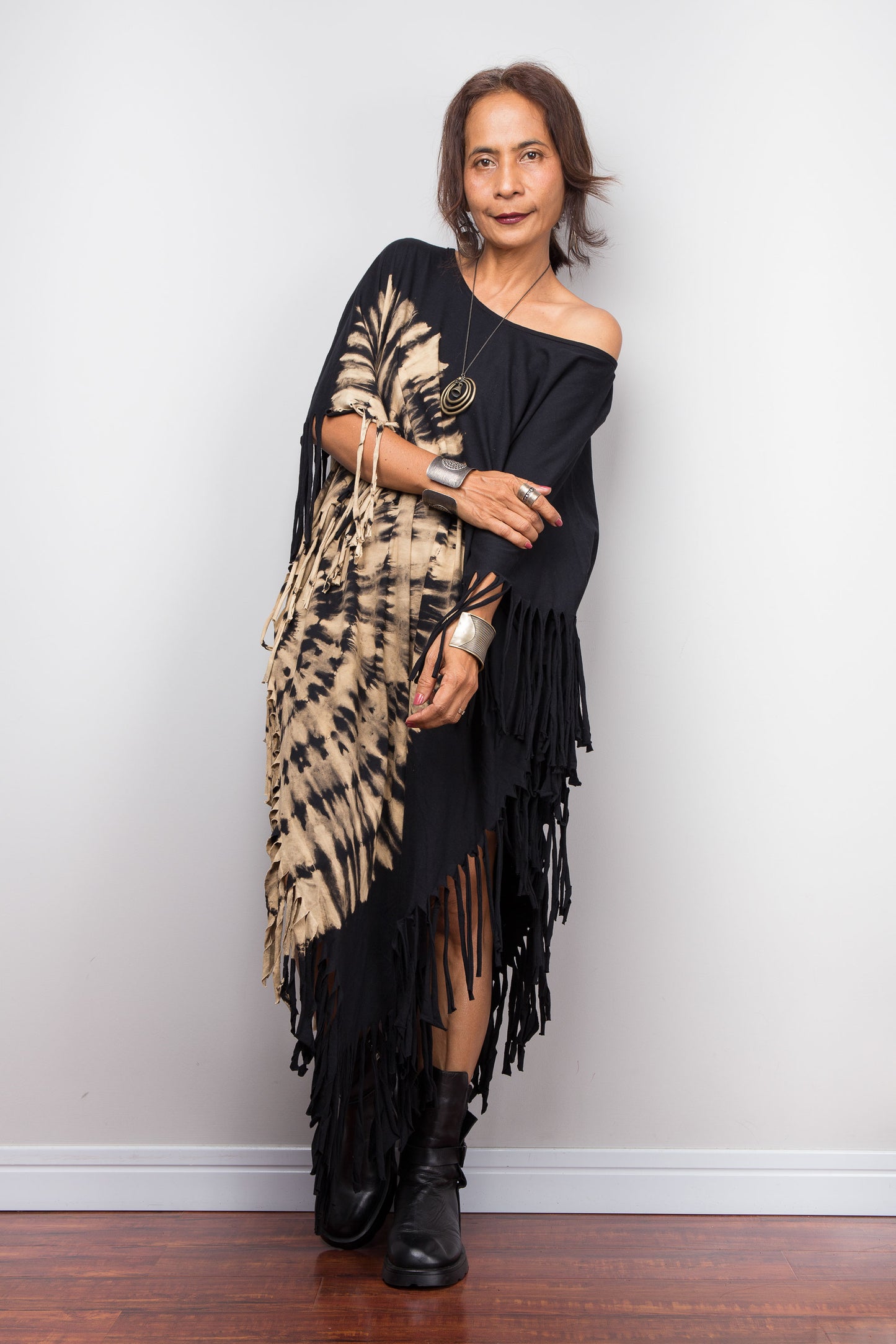 Woman wearing a black off-shoulder reverse tie dye poncho dress with a beige and brown pattern and black boots against a white wall.