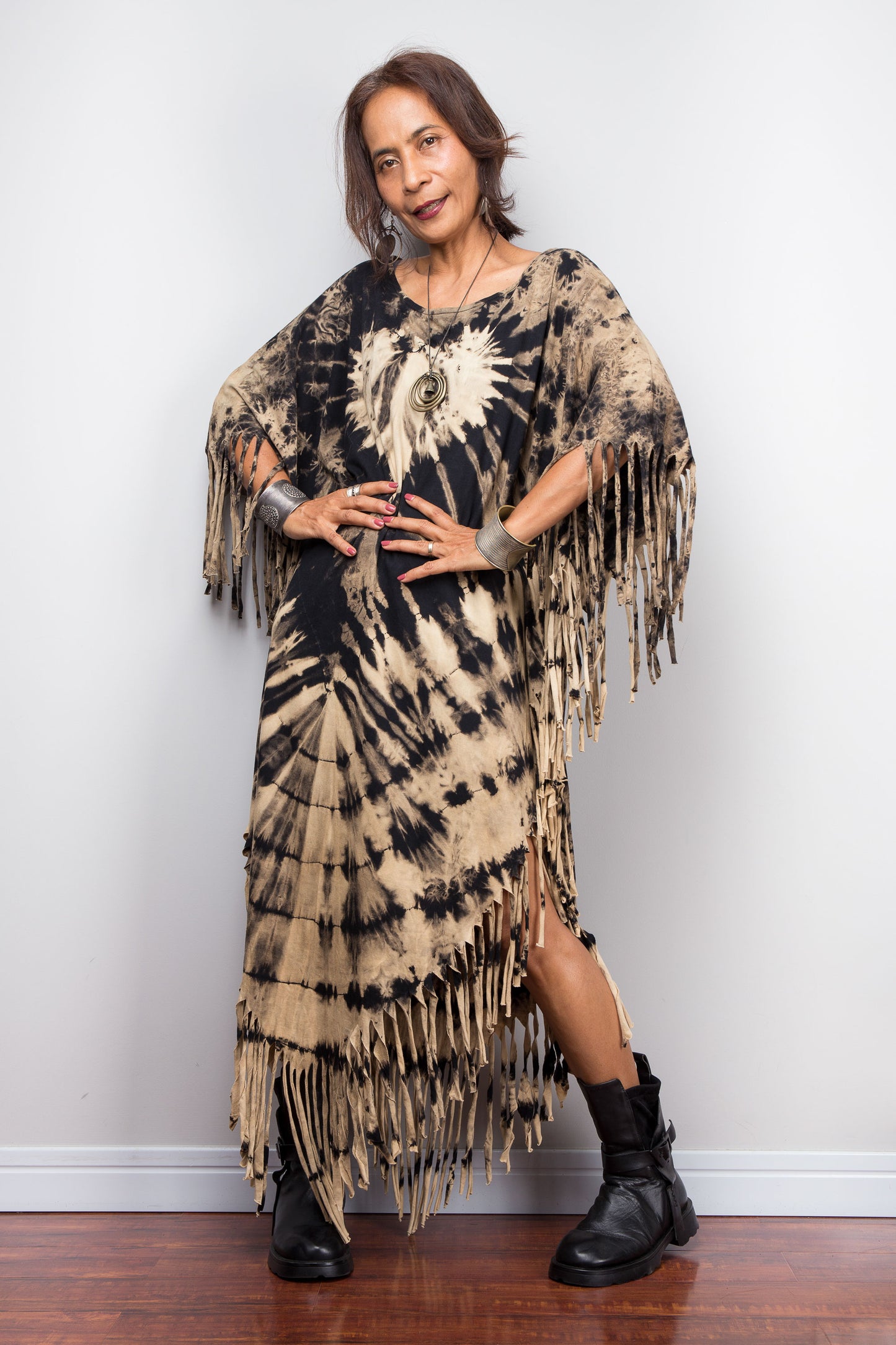Woman wearing a tie-dye dress with fringe and black boots against a white wall.