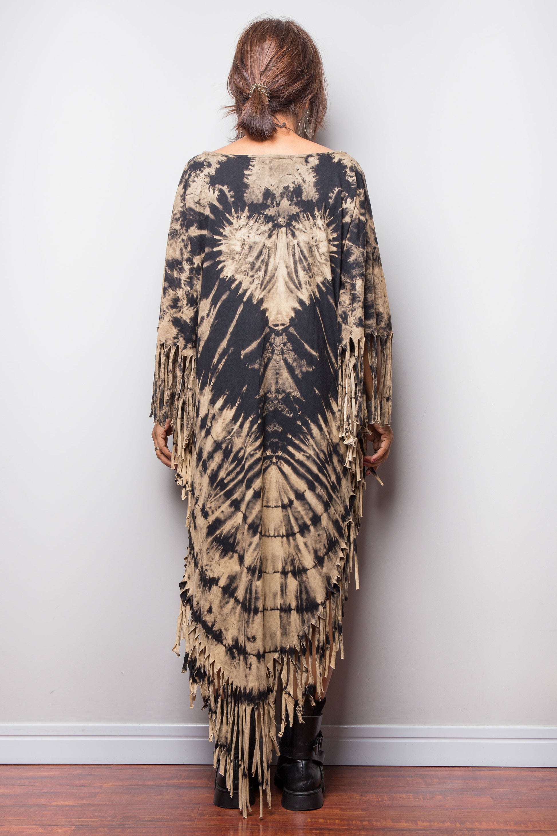 Person wearing a tie-dye dress with fringe details against a plain wall.