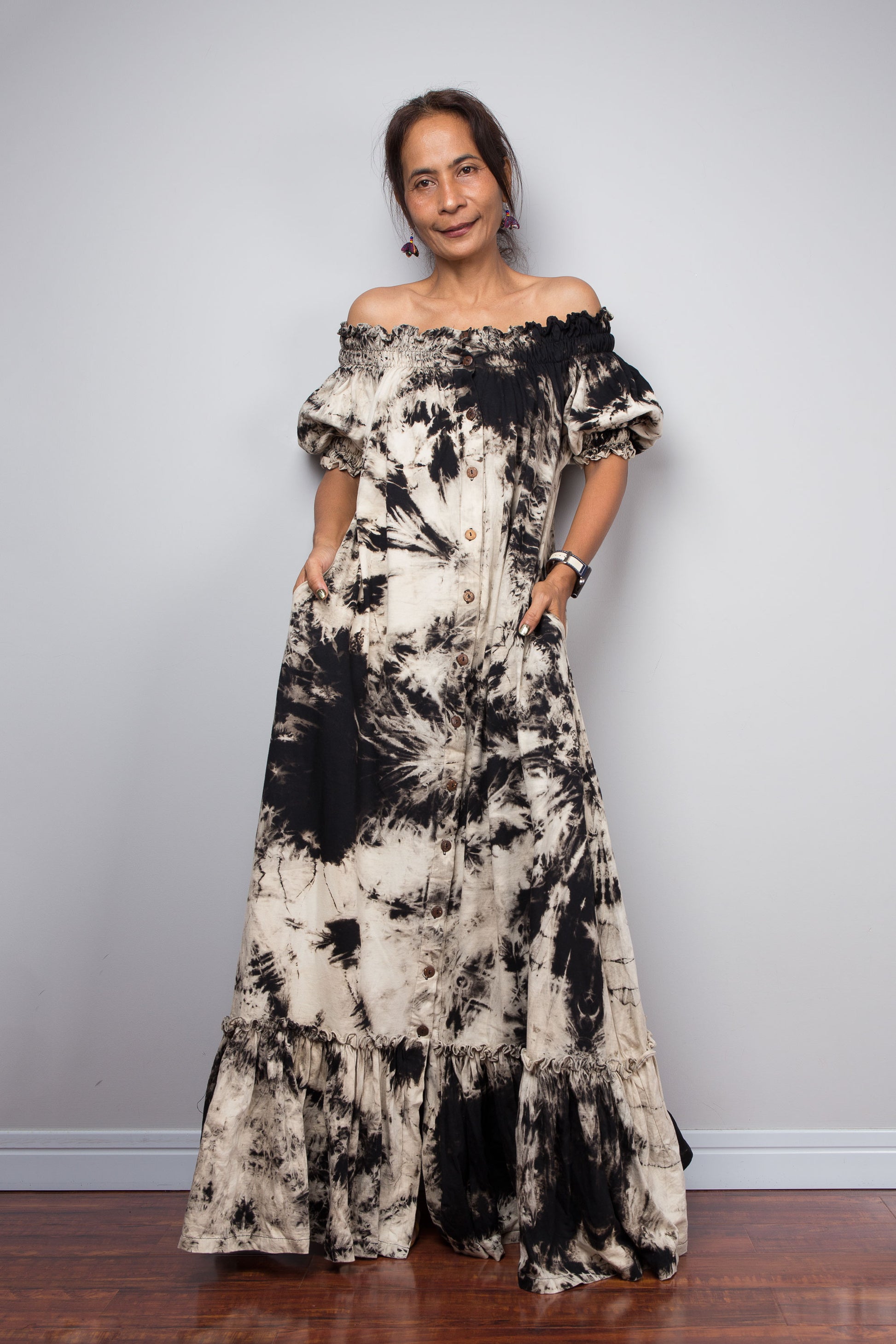 Front view of a model wearing the Nuichan off-the-shoulder reverse tie dye maxi shirt dress in black and sand cotton, showcasing the elasticated smocked neckline worn off the shoulder, full button-down front, and floor-length silhouette