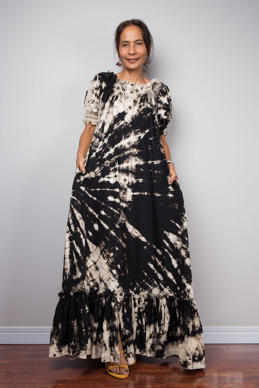 Front-facing view of a model wearing the Nuichan black and sand reverse tie dye off-shoulder maxi shirt dress with hands in pockets, showcasing the bold starburst reverse tie dye pattern, two-way smocked neckline worn as a gathered scoop neck, rustic button-down front, puff sleeves with smocked cuffs, and tiered ruffle hem with sand fade