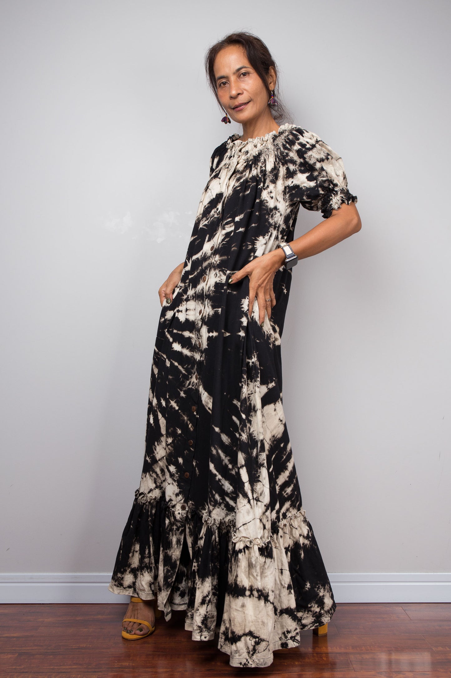 Three quarter view of a model wearing the Nuichan black and sand reverse tie dye off-shoulder maxi shirt dress with hands in pockets, showcasing the bold starburst reverse tie dye pattern, two-way smocked neckline worn as a gathered scoop neck, rustic button-down front, puff sleeves with smocked cuffs, and tiered ruffle hem with sand fade