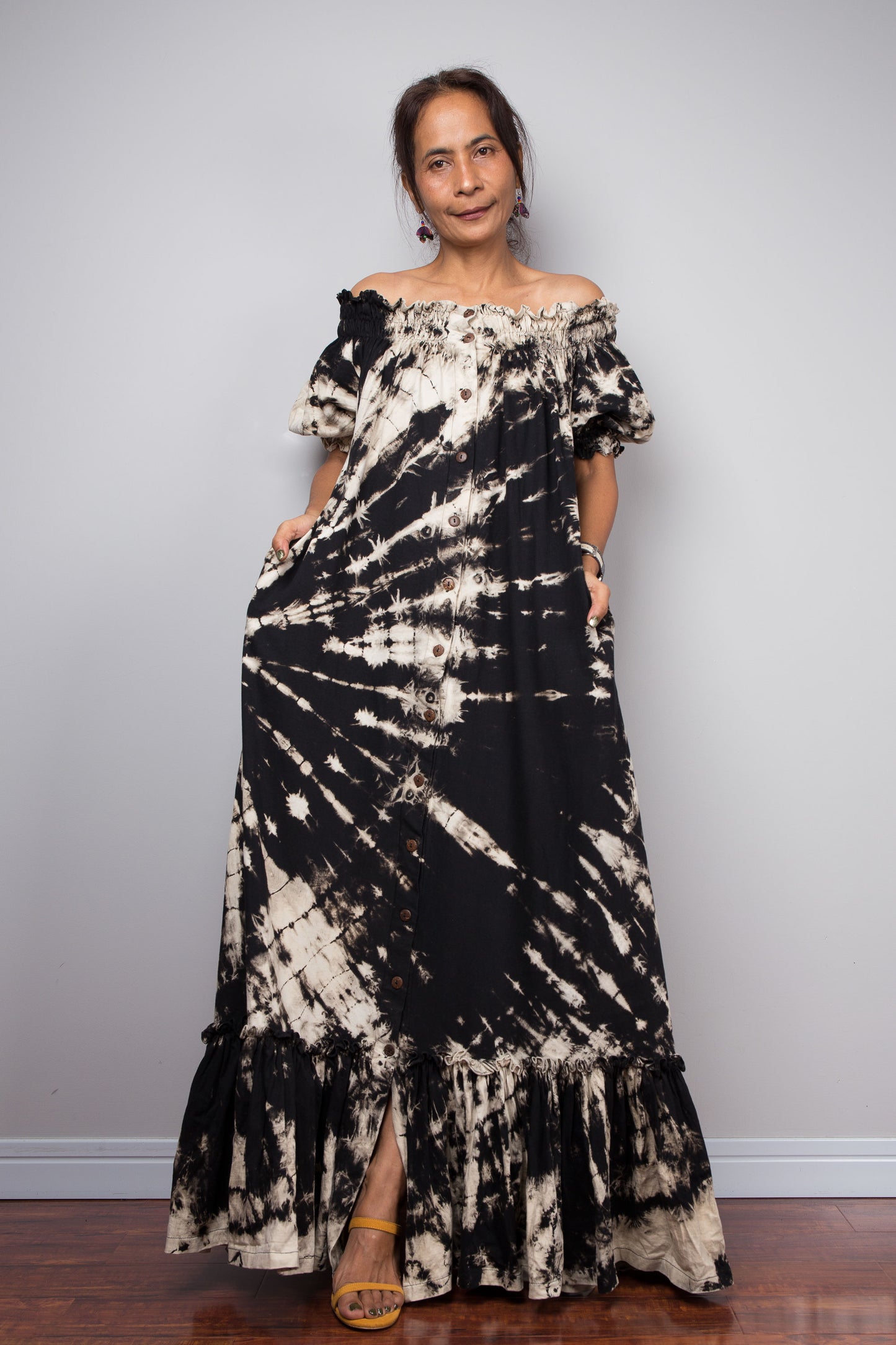 Front-facing view of a model wearing the Nuichan black reverse tie dye off-shoulder maxi shirt dress with hands in pockets, showcasing the bold starburst reverse tie dye pattern, two-way smocked neckline worn as a gathered scoop neck, rustic button-down front, puff sleeves with smocked cuffs, and tiered ruffle hem with sand fade