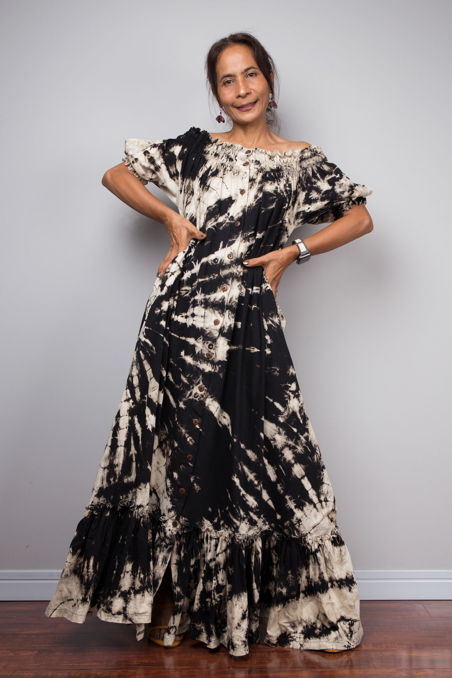 Front-facing view of a model wearing the Nuichan black and sand reverse tie dye off-shoulder maxi shirt dress with hands on hips, showcasing the bold starburst reverse tie dye pattern, two-way smocked neckline worn as a gathered scoop neck, rustic button-down front, puff sleeves with smocked cuffs, and tiered ruffle hem with sand fade