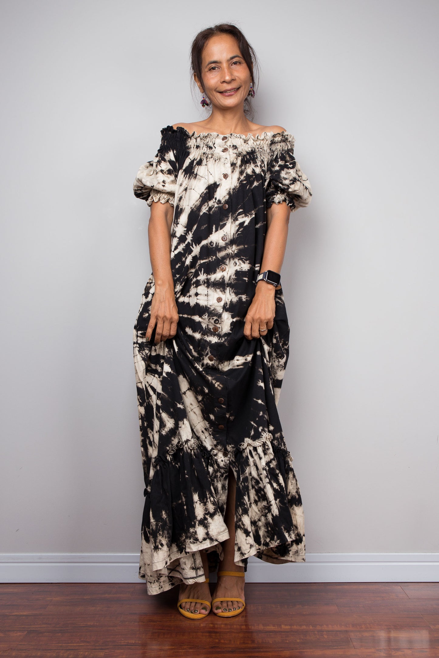 Front-facing view of a model wearing the Nuichan black and sand reverse tie dye off-shoulder maxi shirt dress with hands pulling up the lower part of the dress , showcasing the bold starburst reverse tie dye pattern, two-way smocked neckline worn as a gathered scoop neck, rustic button-down front, puff sleeves with smocked cuffs, and tiered ruffle hem with sand fade