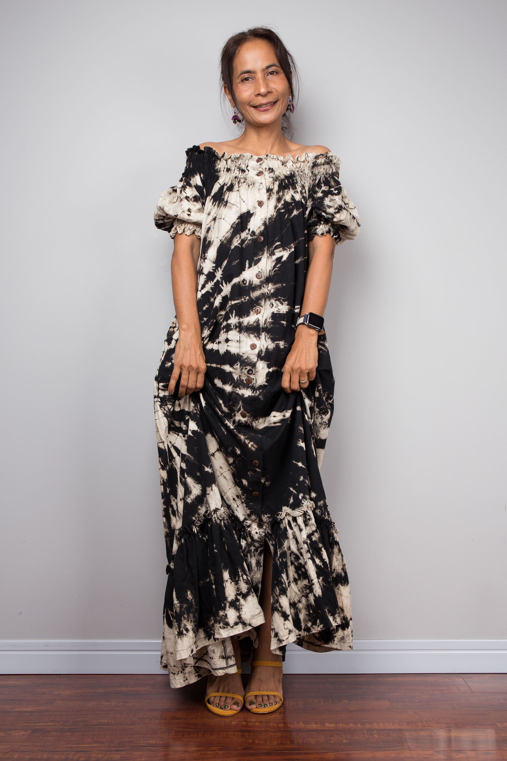 Front-facing view of a model wearing the Nuichan black and sand reverse tie dye off-shoulder maxi shirt dress with hands pulling up the lower part of the dress , showcasing the bold starburst reverse tie dye pattern, two-way smocked neckline worn as a gathered scoop neck, rustic button-down front, puff sleeves with smocked cuffs, and tiered ruffle hem with sand fade