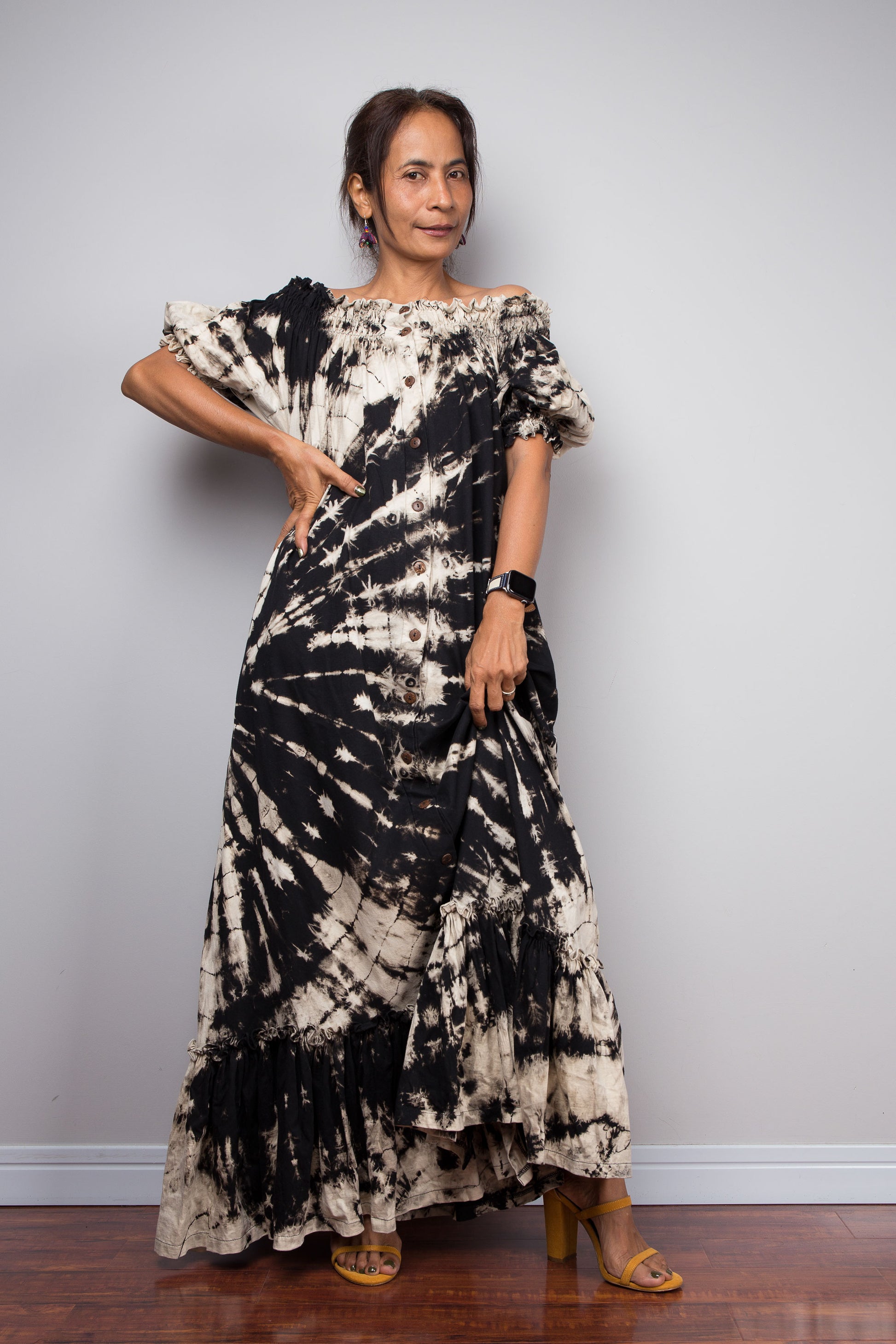 Frontal view of a model wearing the Nuichan black and sand reverse tie dye off-shoulder maxi shirt dress with hand on hip, showcasing the bold starburst reverse tie dye pattern, two-way smocked neckline worn as a gathered scoop neck, rustic button-down front, puff sleeves with smocked cuffs, and tiered ruffle hem with sand fade
