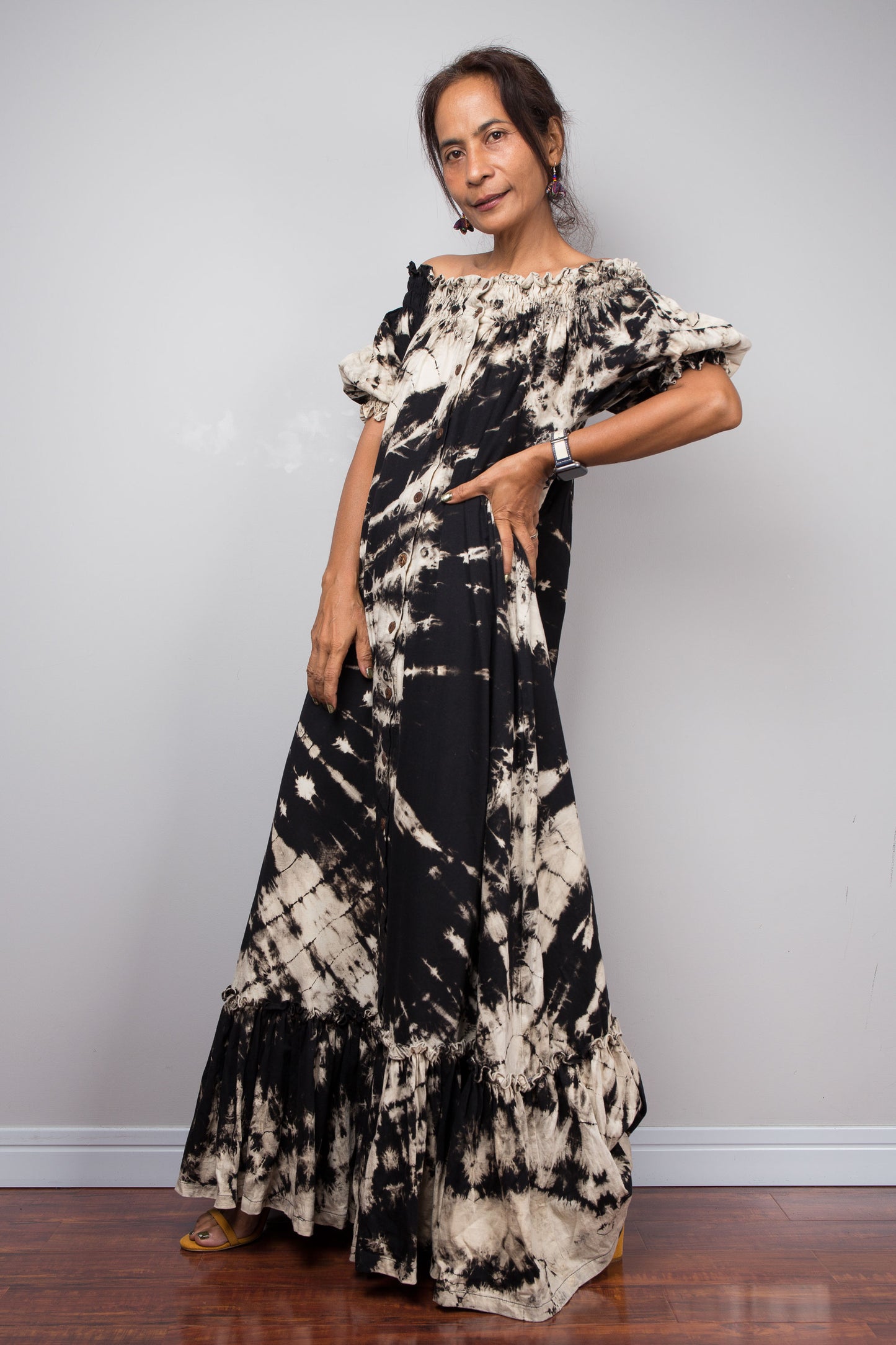 Side view of a model wearing the Nuichan black and sand reverse tie dye off-shoulder maxi shirt dress with hand on the hip, showcasing the bold starburst reverse tie dye pattern, two-way smocked neckline worn as a gathered scoop neck, rustic button-down front, puff sleeves with smocked cuffs, and tiered ruffle hem with sand fade