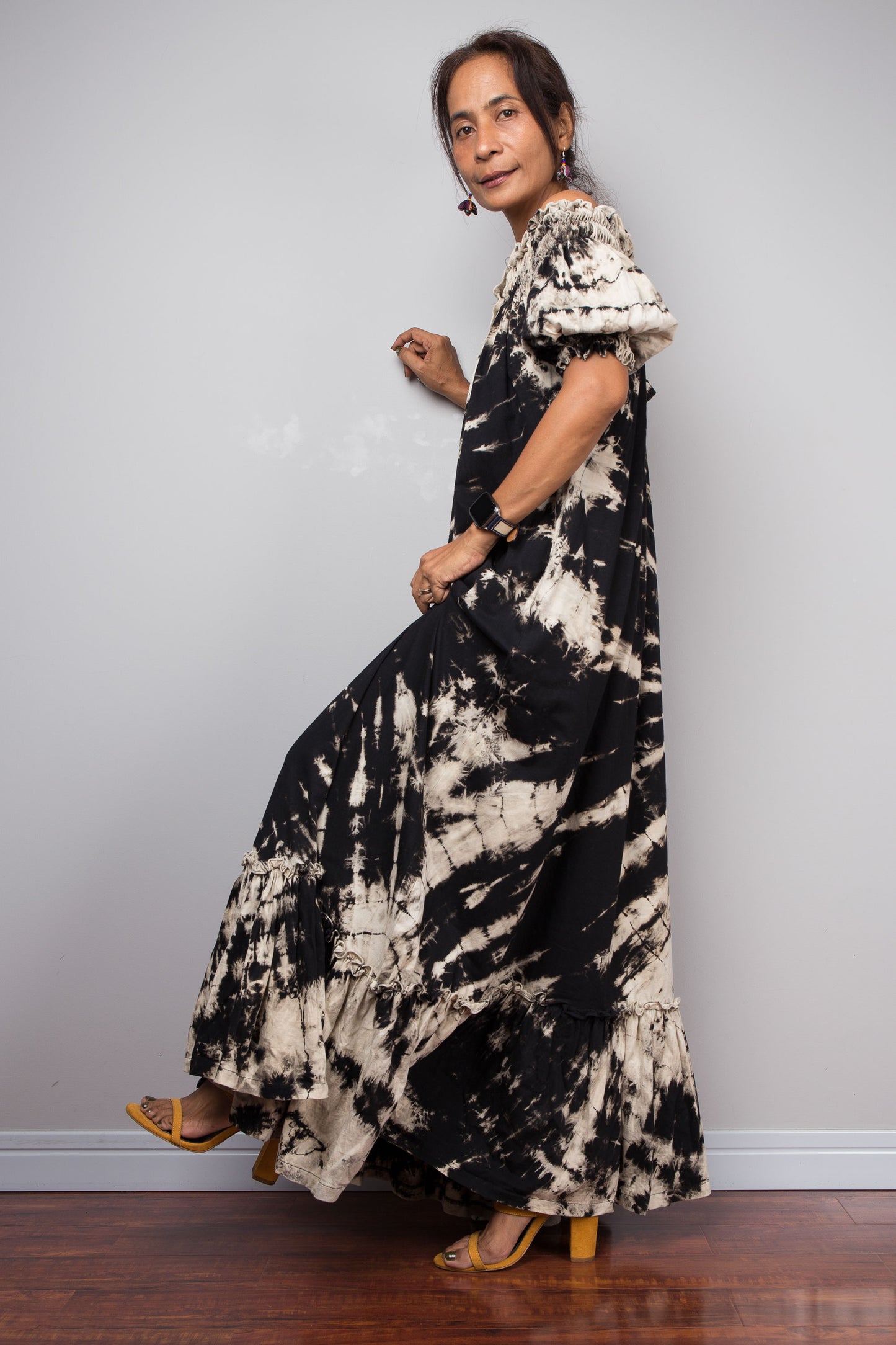 Side view of a model wearing the Nuichan black and sand reverse tie dye off-shoulder maxi shirt dress with one leg lifted, showcasing the bold starburst reverse tie dye pattern, two-way smocked neckline worn as a gathered scoop neck, rustic button-down front, puff sleeves with smocked cuffs, and tiered ruffle hem with sand fade
