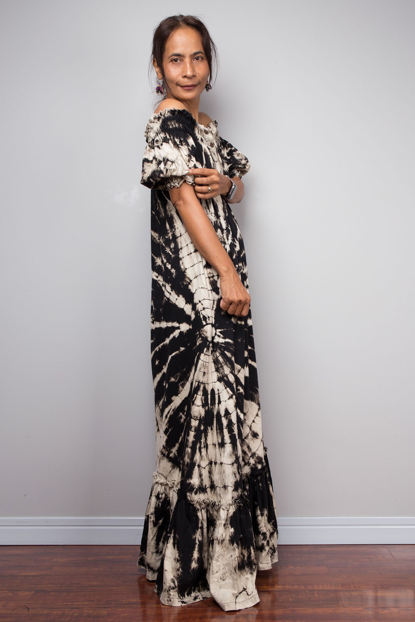 Side view of a model wearing the Nuichan black and sand reverse tie dye off-shoulder maxi shirt dress with hands on the sleeve, showcasing the bold starburst reverse tie dye pattern, two-way smocked neckline worn as a gathered scoop neck, rustic button-down front, puff sleeves with smocked cuffs, and tiered ruffle hem with sand fade