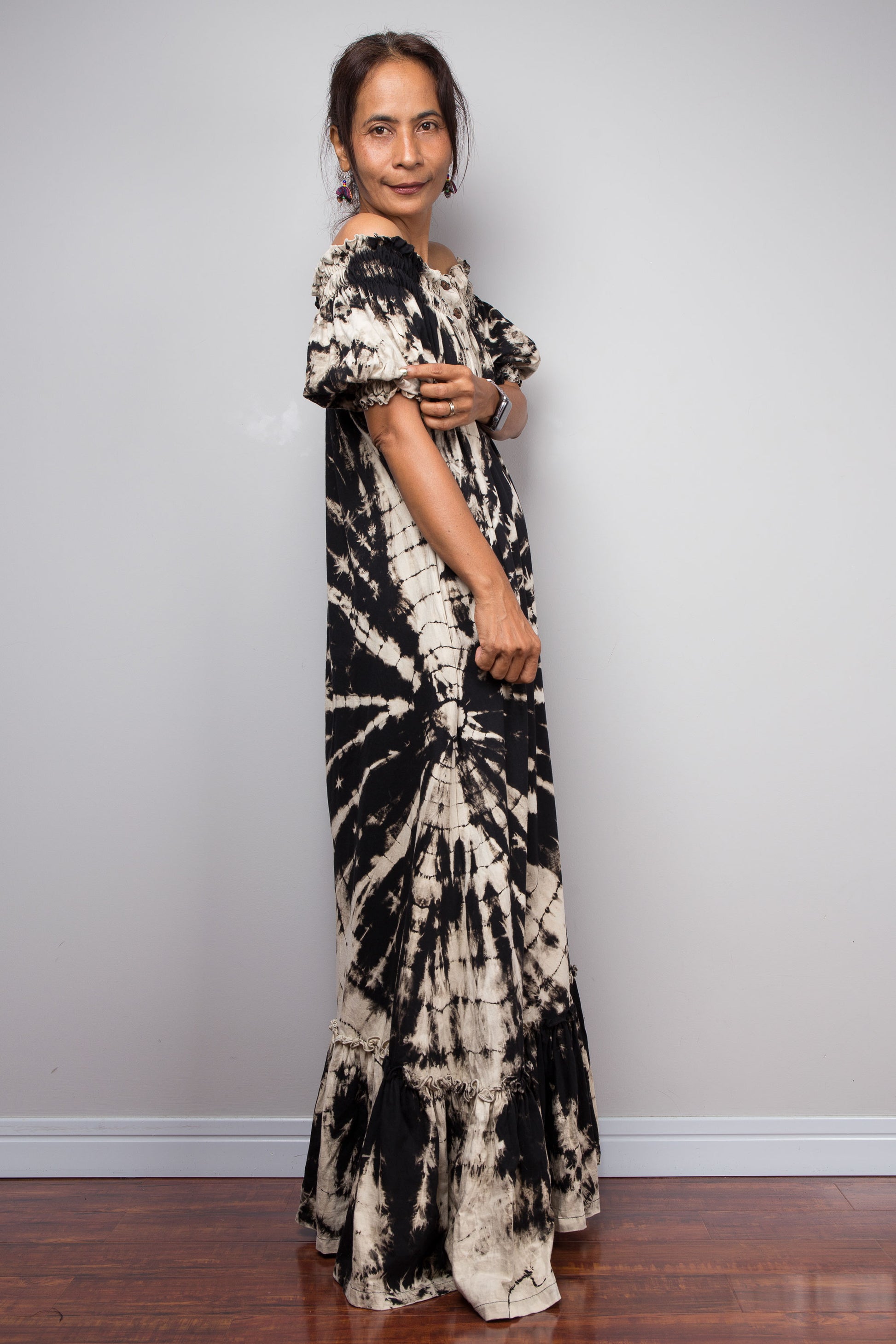 Side view of a model wearing the Nuichan black and sand reverse tie dye off-shoulder maxi shirt dress with hands on the sleeve, showcasing the bold starburst reverse tie dye pattern, two-way smocked neckline worn as a gathered scoop neck, rustic button-down front, puff sleeves with smocked cuffs, and tiered ruffle hem with sand fade