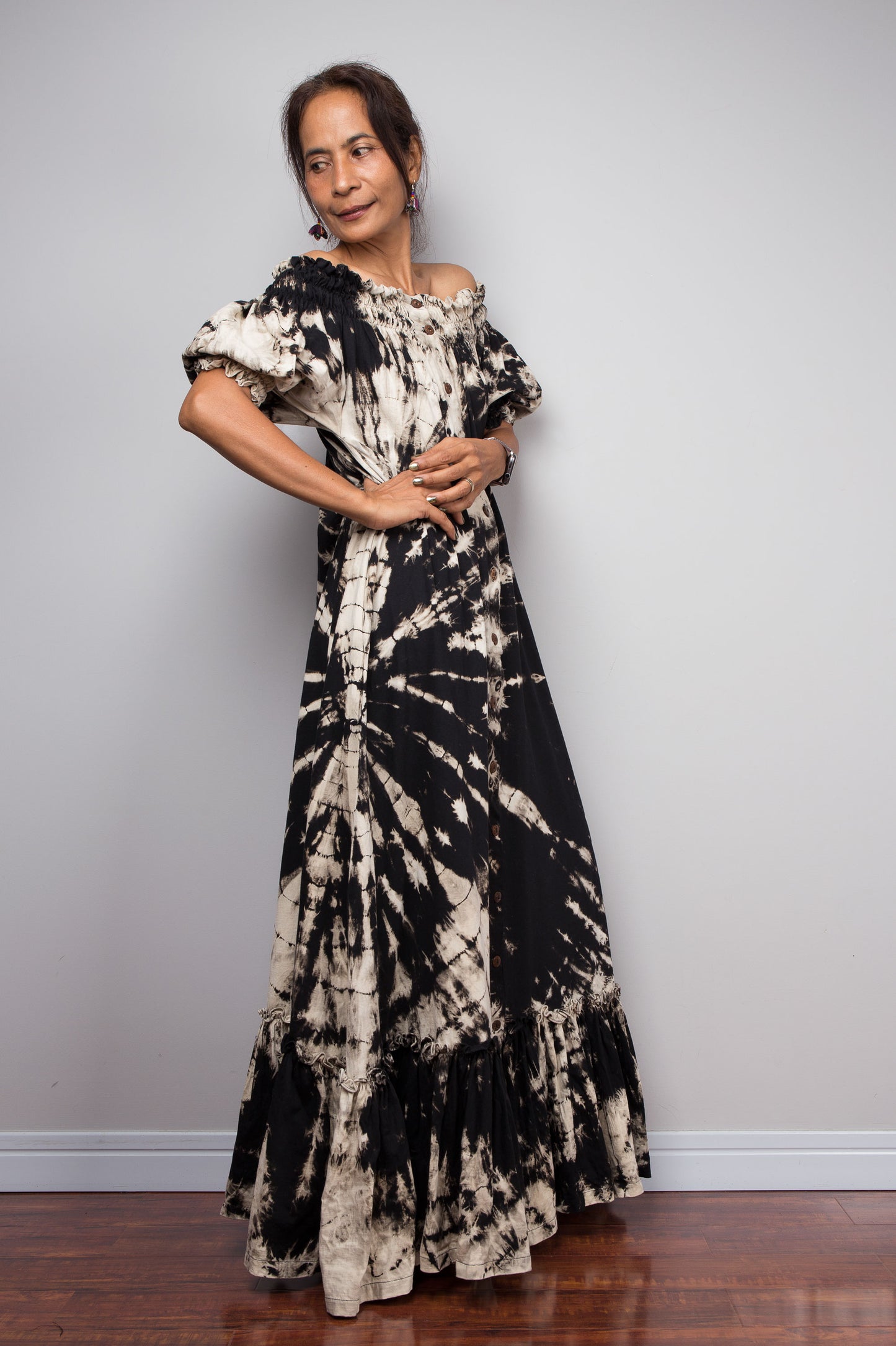 Three quarter back-facing view of a model wearing the Nuichan black and sand reverse tie dye off-shoulder maxi shirt dress with hands on hips, showcasing the bold starburst reverse tie dye pattern, two-way smocked neckline worn as a gathered scoop neck, rustic button-down front, puff sleeves with smocked cuffs, and tiered ruffle hem with sand fade
