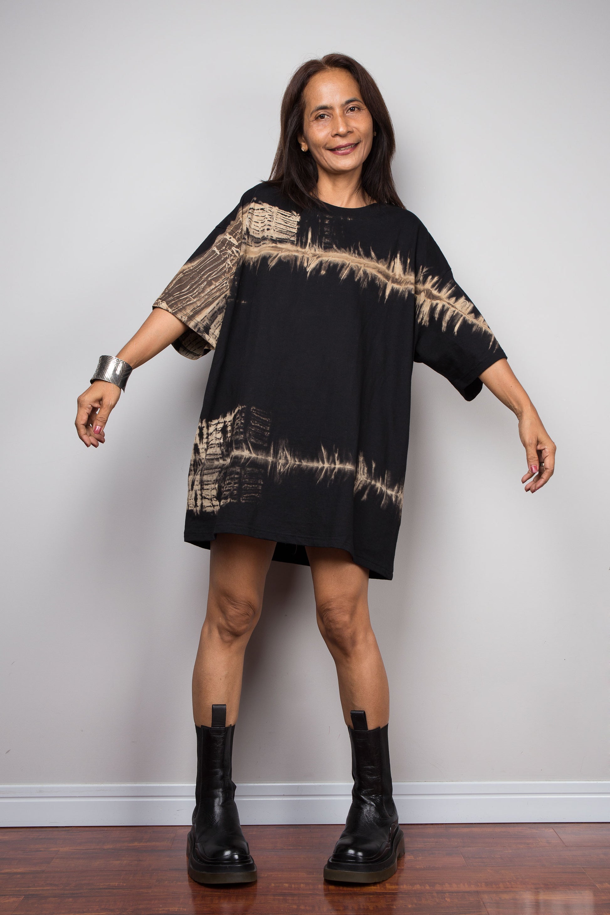 A woman modeling a black oversized short-sleeve tee with beige tie-dye patterns, paired with black leather boots, standing against a light gray wall.