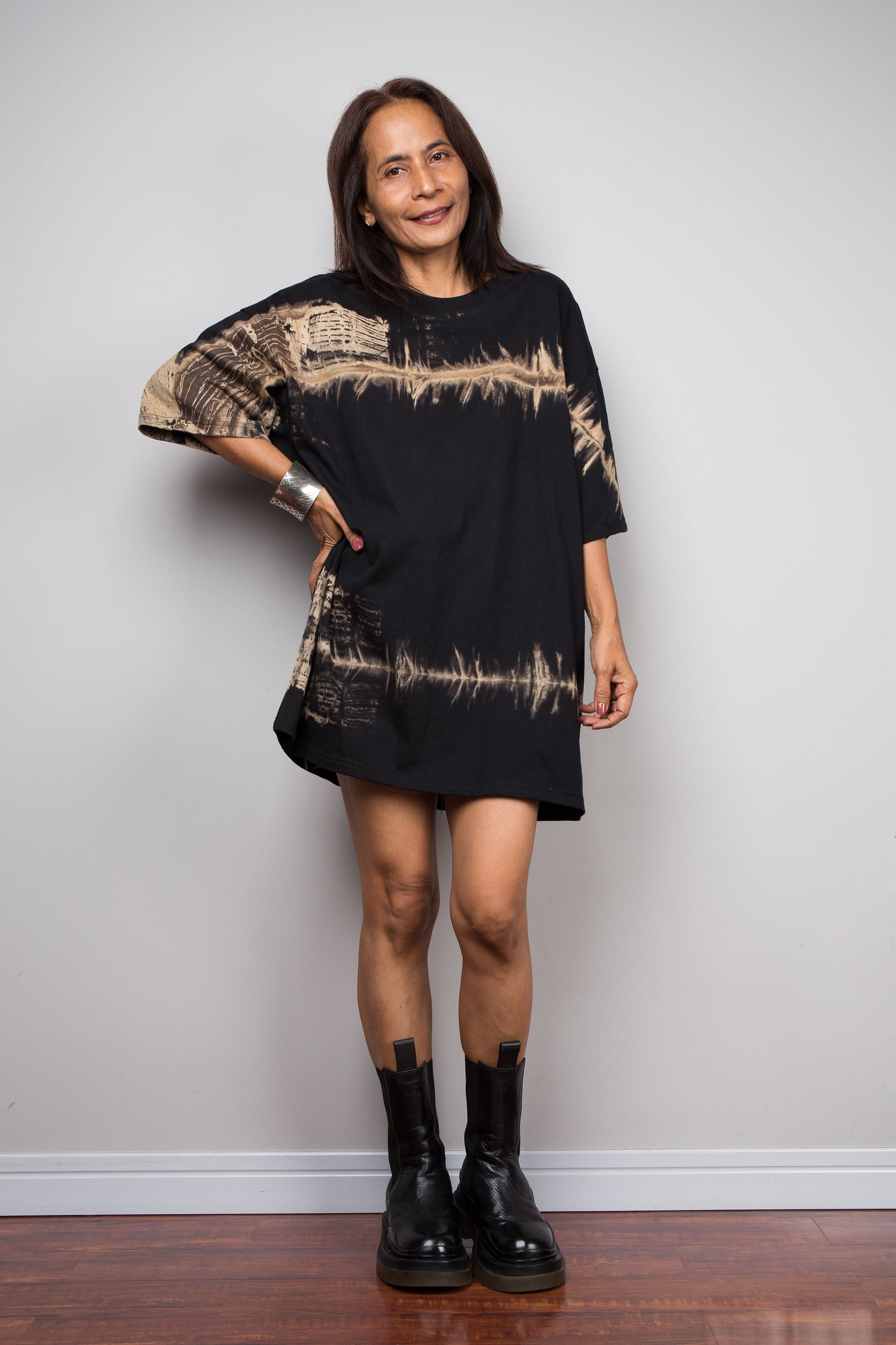 A woman modeling a Nuichan black oversized short-sleeve t shirt with beige soundwave-like graphic tie dye patterns, paired with black ankle boots, standing against a light gray wall.