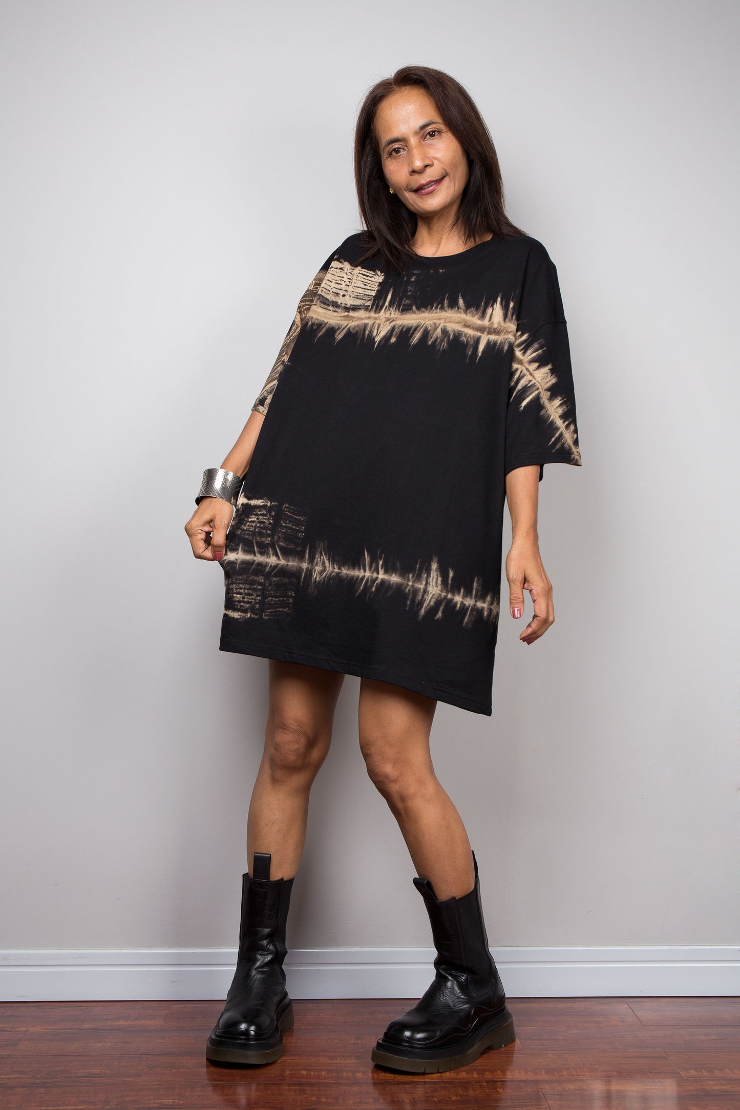 A woman modeling a Nuichan black oversized short-sleeve t shirt with beige soundwave-like reverse tie dye patterns, paired with black leather ankle boots, standing against a light gray wall.