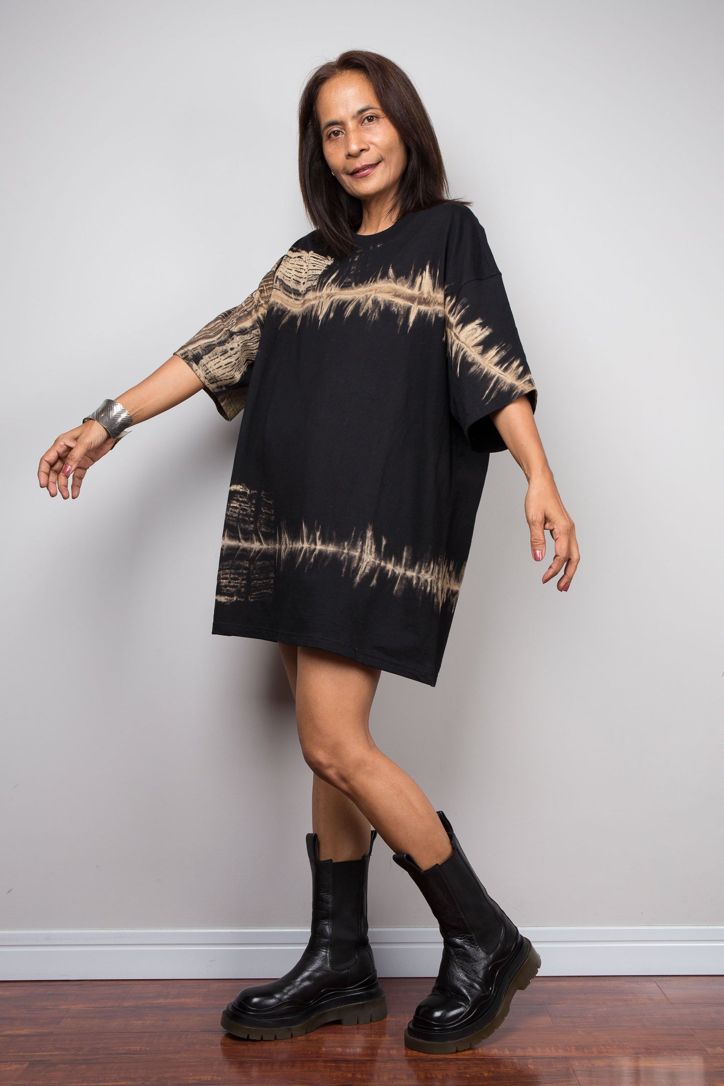 A woman modeling an oversized black t shirt dress with beige tie-dye patterns on the sleeves and hem, paired with black leather ankle boots, standing against a light gray wall.