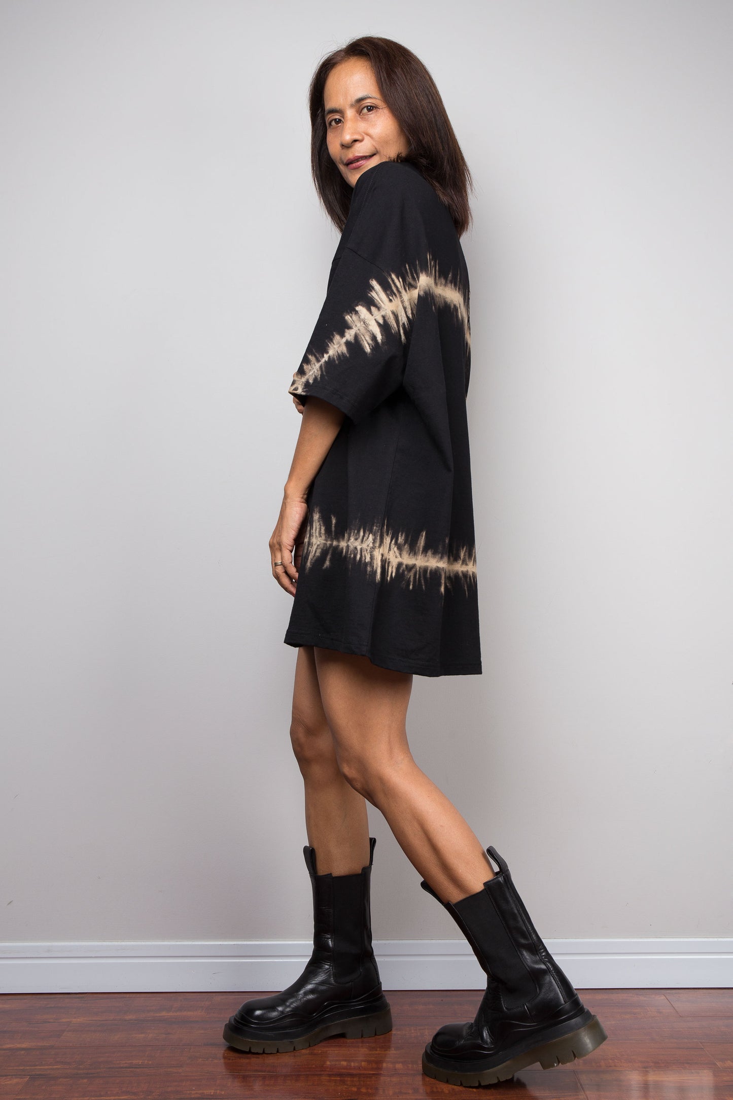 A woman modeling a Nuichan black short-sleeve t shirt dress with beige reverse tie-dye soundwave patterns on the sleeves and hem, paired with black leather boots, standing against a light gray wall.