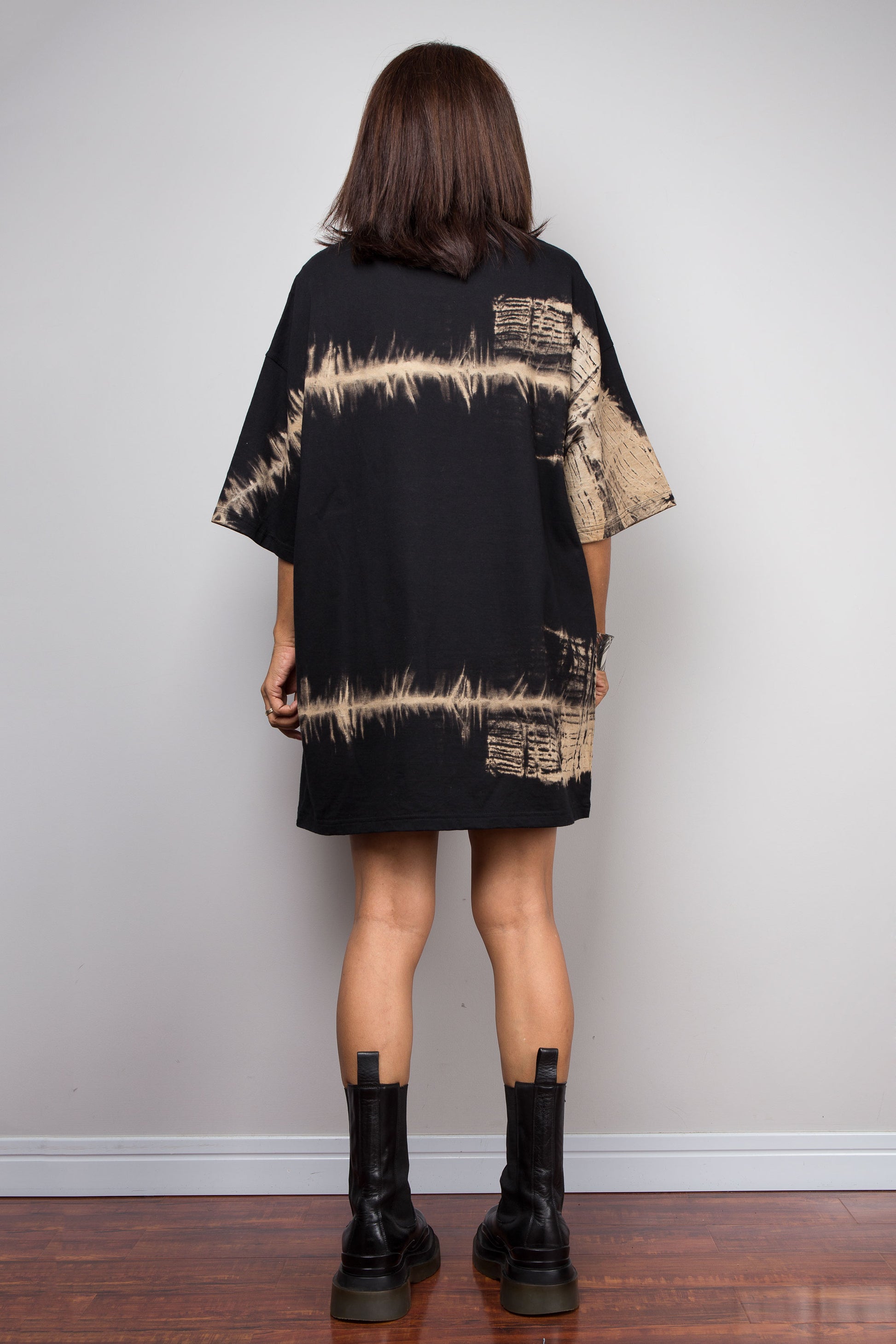 A Nuichan  model viewed from behind wearing a black oversized short-sleeve t-shirt dress with a beige tie-dye pattern, paired with black leather ankle boots, standing against a plain light gray wall.