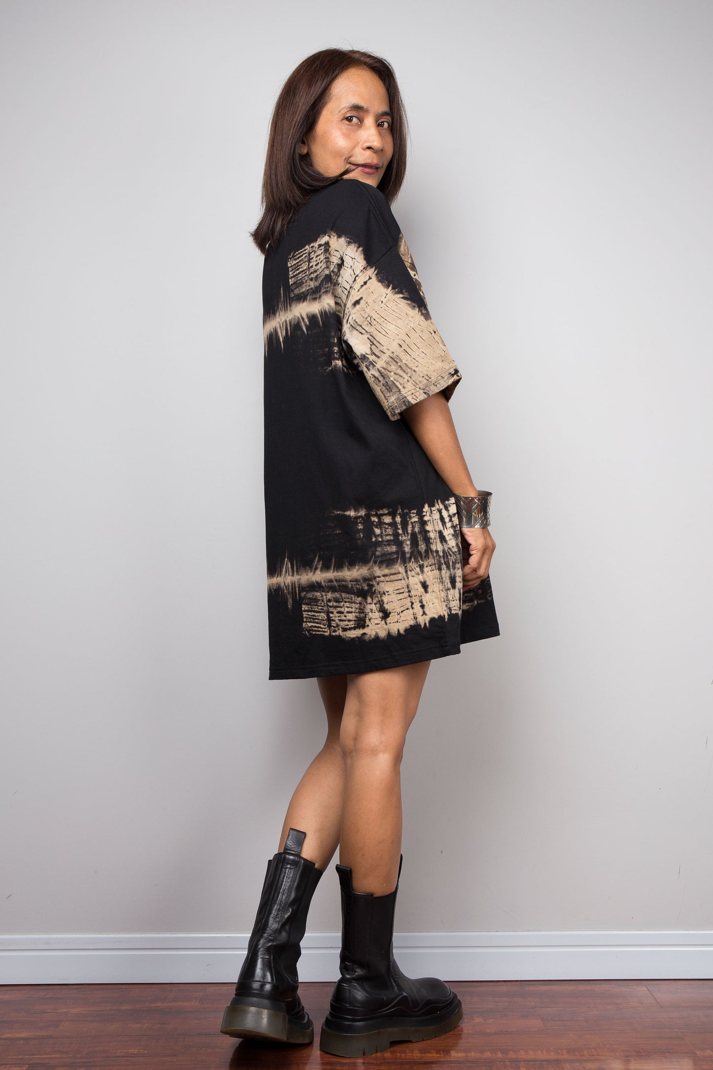 A woman modeling a black oversized t-shirt dress with beige abstract Nuichan tie-dye patterns, paired with black leather ankle boots, standing against a light gray wall.