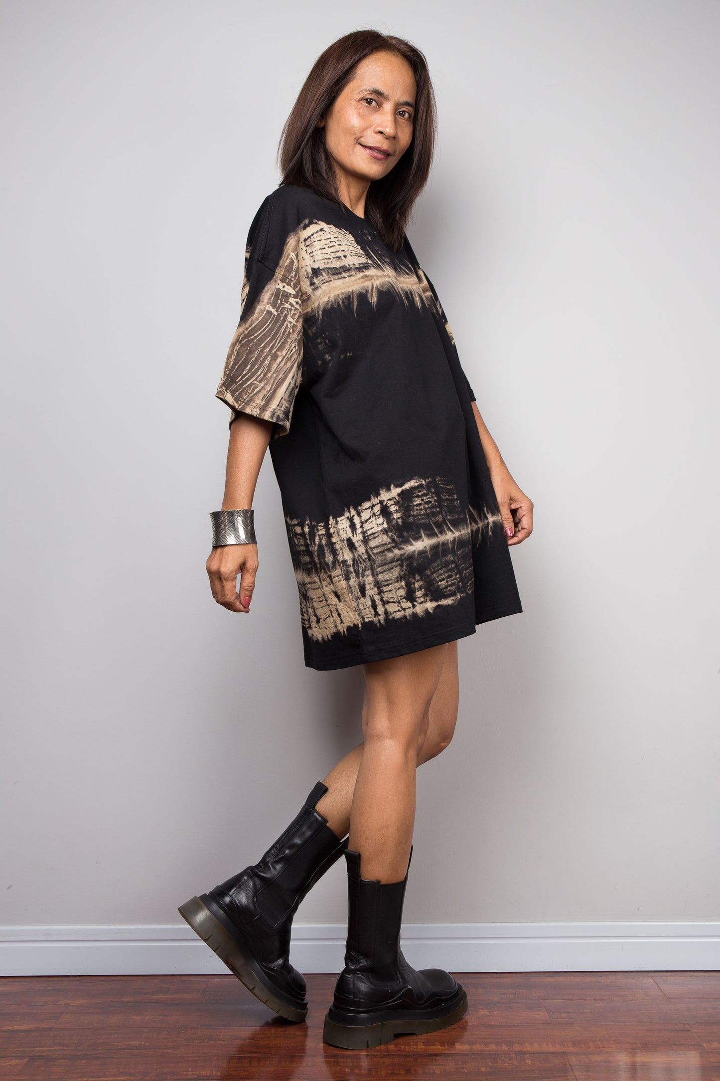 A woman modeling a black short-sleeve dress with beige abstract brushstroke patterns on the sleeves and hem, paired with black boots, standing against a light gray wall.