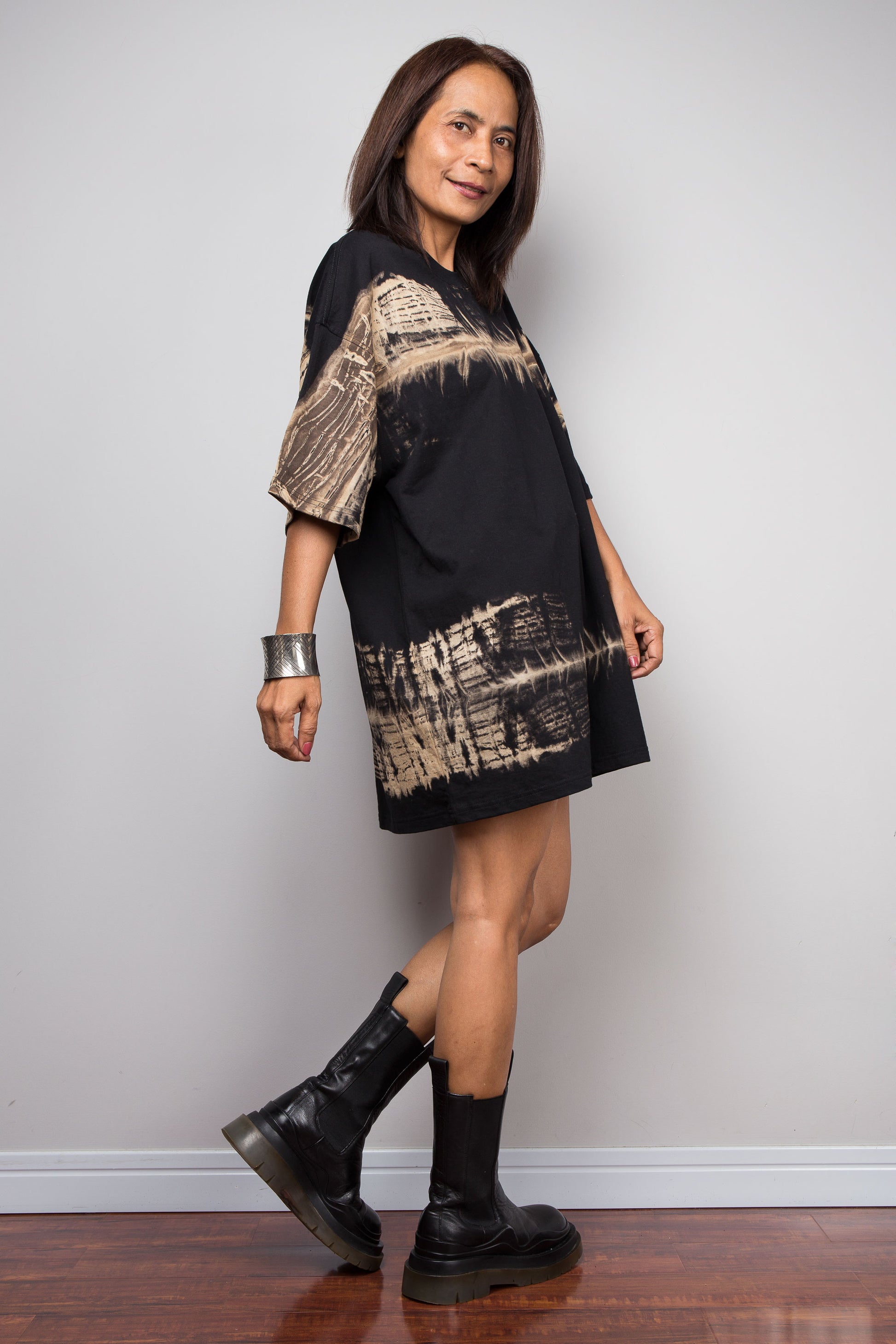 A woman modeling a black short-sleeve dress with beige abstract brushstroke patterns on the sleeves and hem, paired with black boots, standing against a light gray wall.