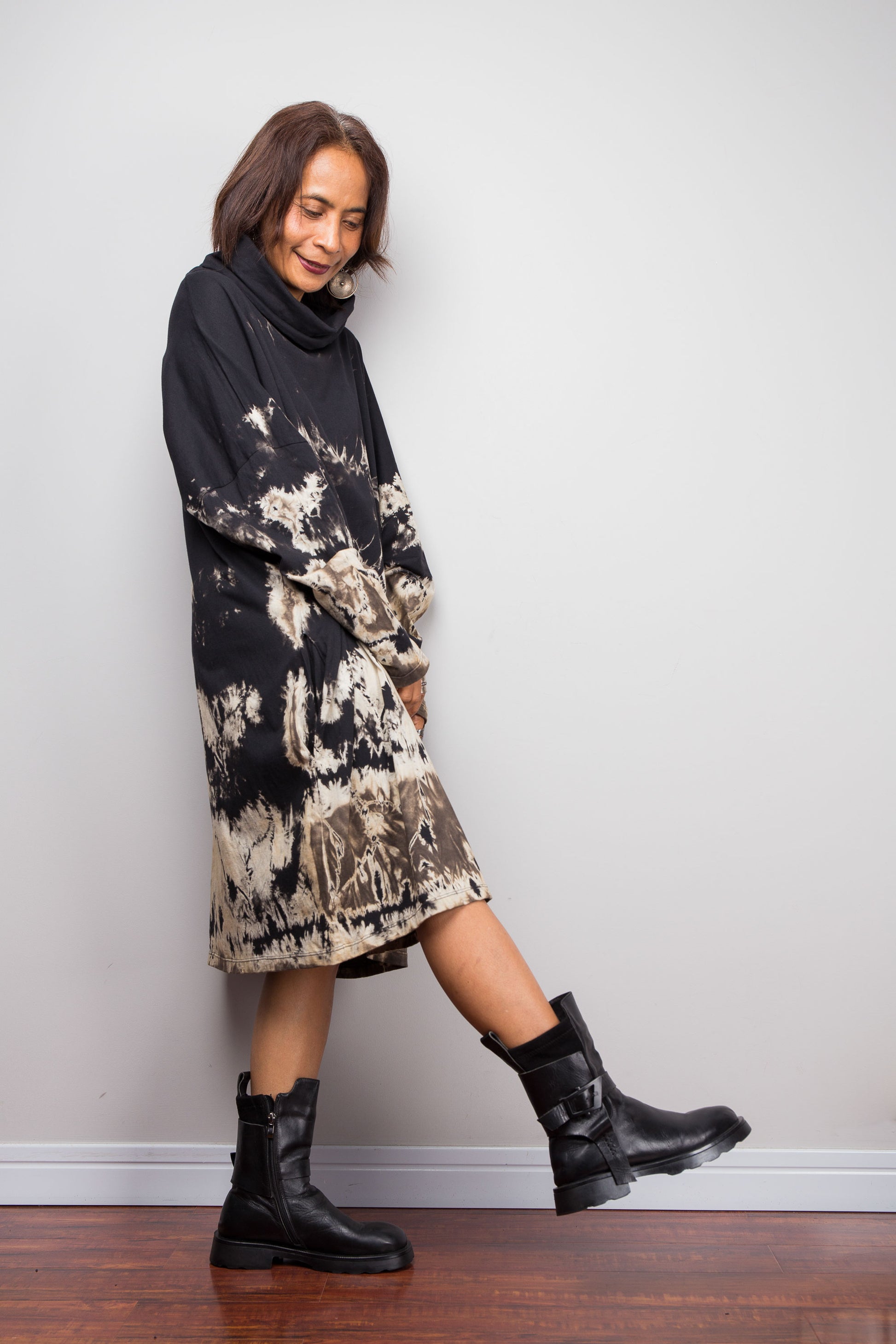 Petite women wearing an Oversized Nuichan reverse tie-dye tunic dress featuring a funnel cowl neck and abstract sand and black patterns.  Side view with one leg stretched out to show the width at the bottom of the tunic