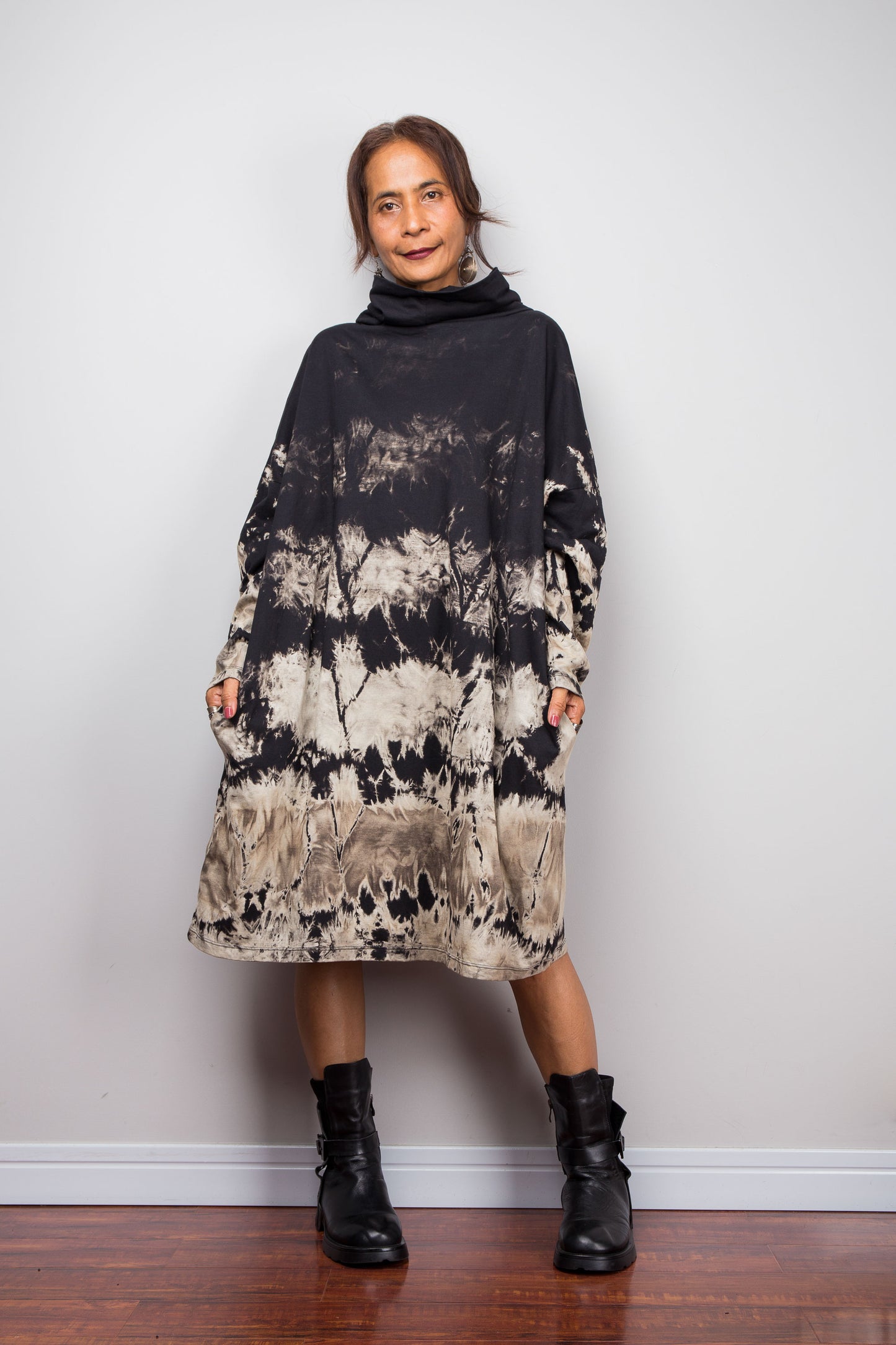 Full front view of a model wearing Nuichan's oversized reverse tie dye tunic dress with a high funnel cowl neck, pockets and horizontal abstract sand-colored patterns.