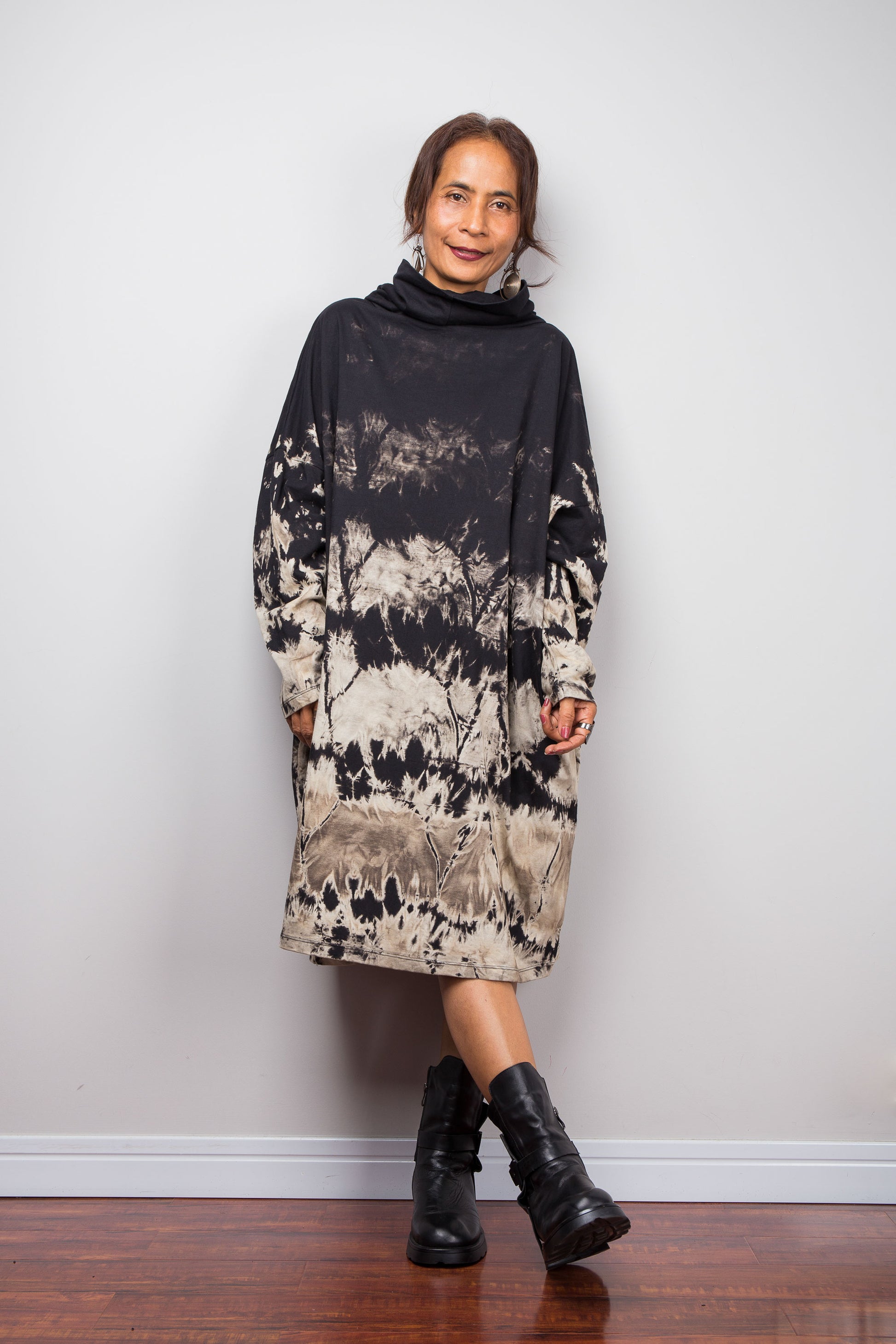 Full front view of a model wearing Nuichan's oversized reverse tie dye tunic dress with a high funnel cowl neck, long sleeves and horizontal abstract sand-colored patterns.