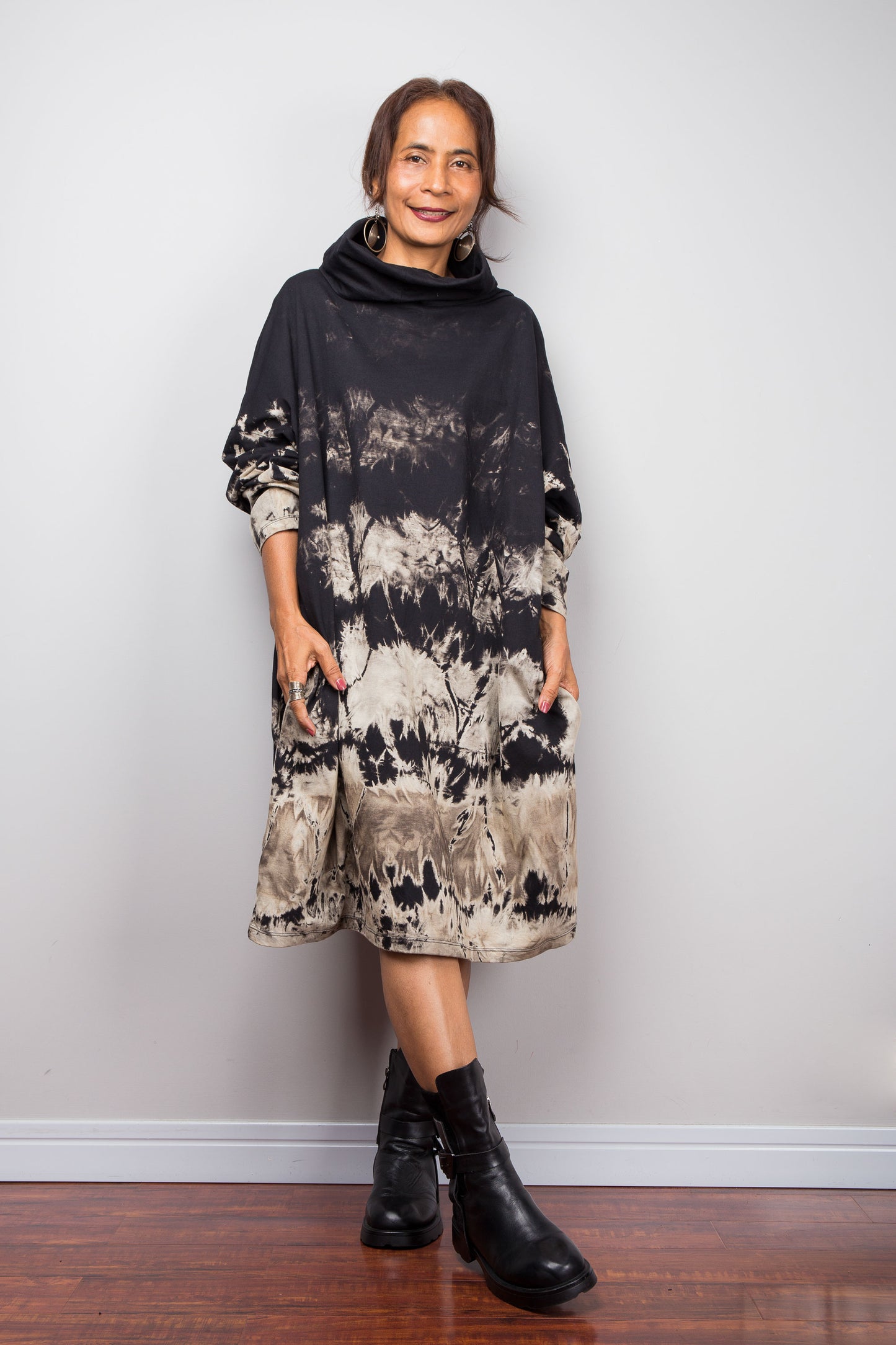 Full front view of a model wearing Nuichan's oversized reverse tie dye tunic dress with pockets and long sleeves, a high funnel cowl neck and horizontal abstract sand-colored patterns.