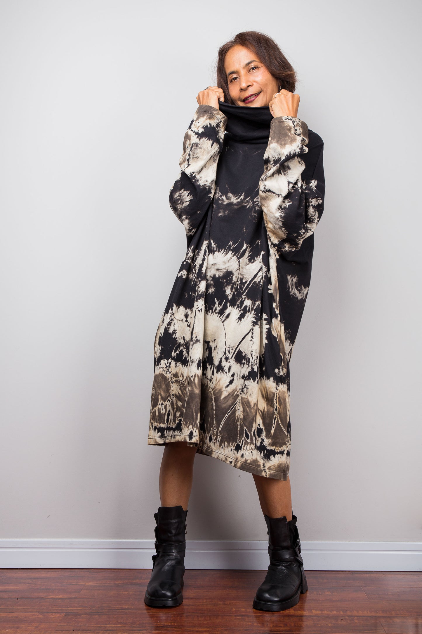 Petite women wearing an Oversized Nuichan reverse tie-dye tunic dress featuring a funnel cowl neck and abstract sand and black patterns.  Frontal view with both hands holding up the funnel neck