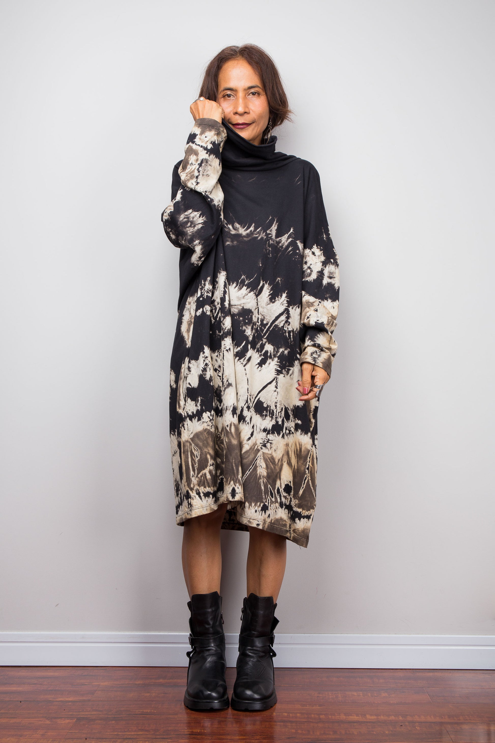 Petite women wearing an Oversized Nuichan reverse tie-dye tunic dress featuring a funnel cowl neck and abstract sand and black patterns.  Frontal view with  one hand holding up the cowl neck