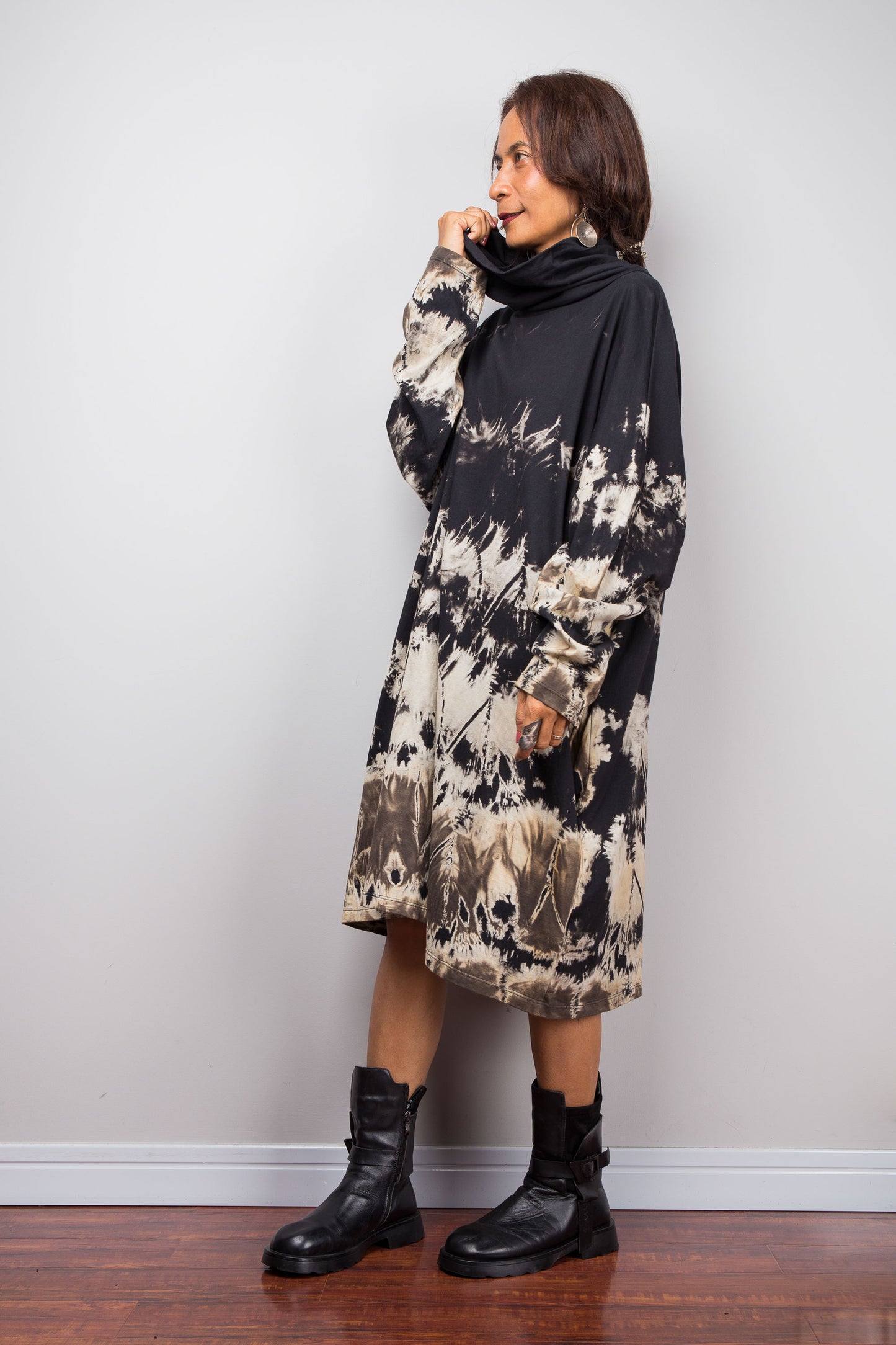 Petite women wearing an Oversized Nuichan reverse tie-dye tunic dress featuring a funnel cowl neck and abstract sand and black patterns.  Side view with one hand holding up the cowl neck
