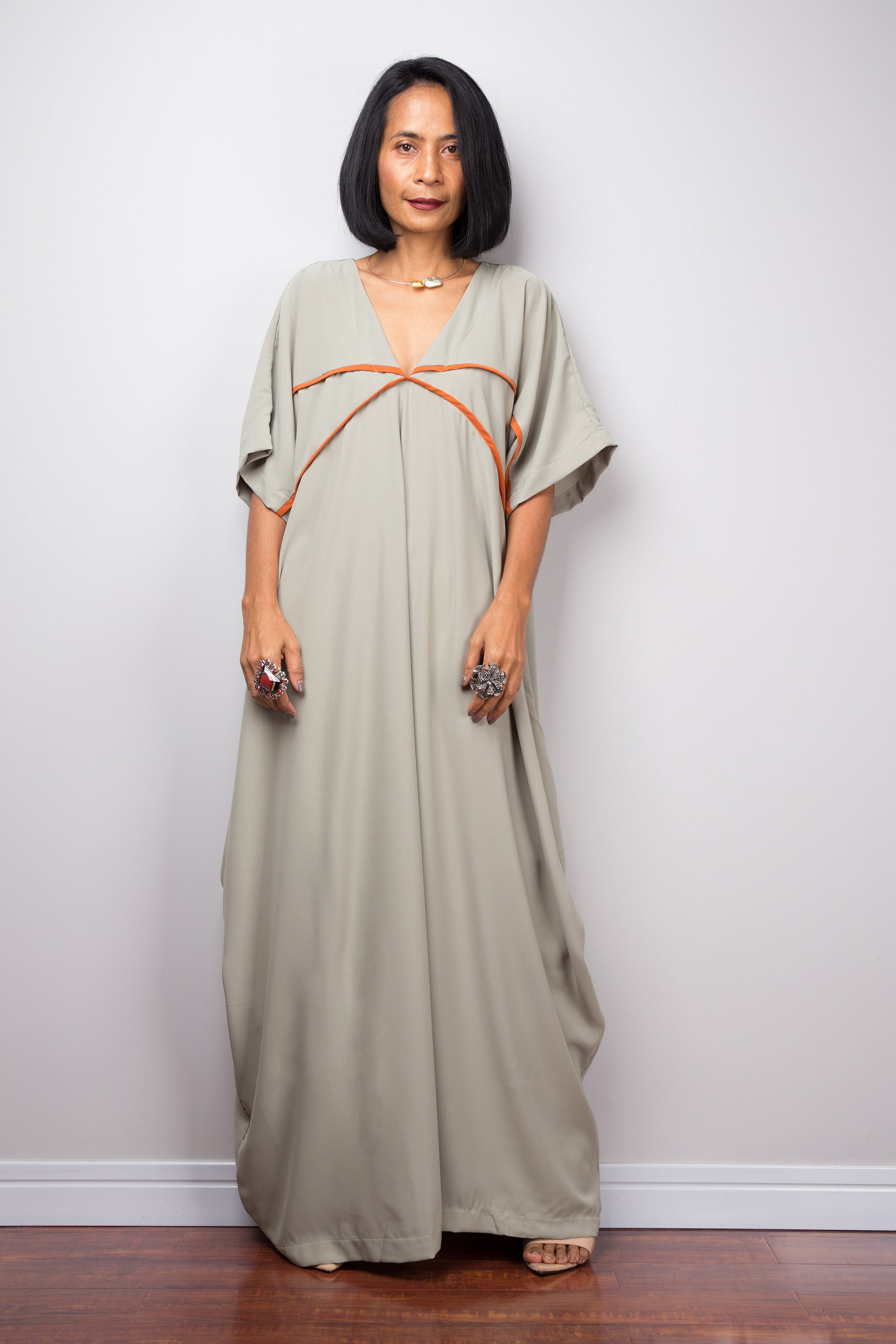 Woman wearing a sage kaftan maxi dress with white crisscross trim, standing against a gray wall on a wooden floor, featuring wide sleeves.