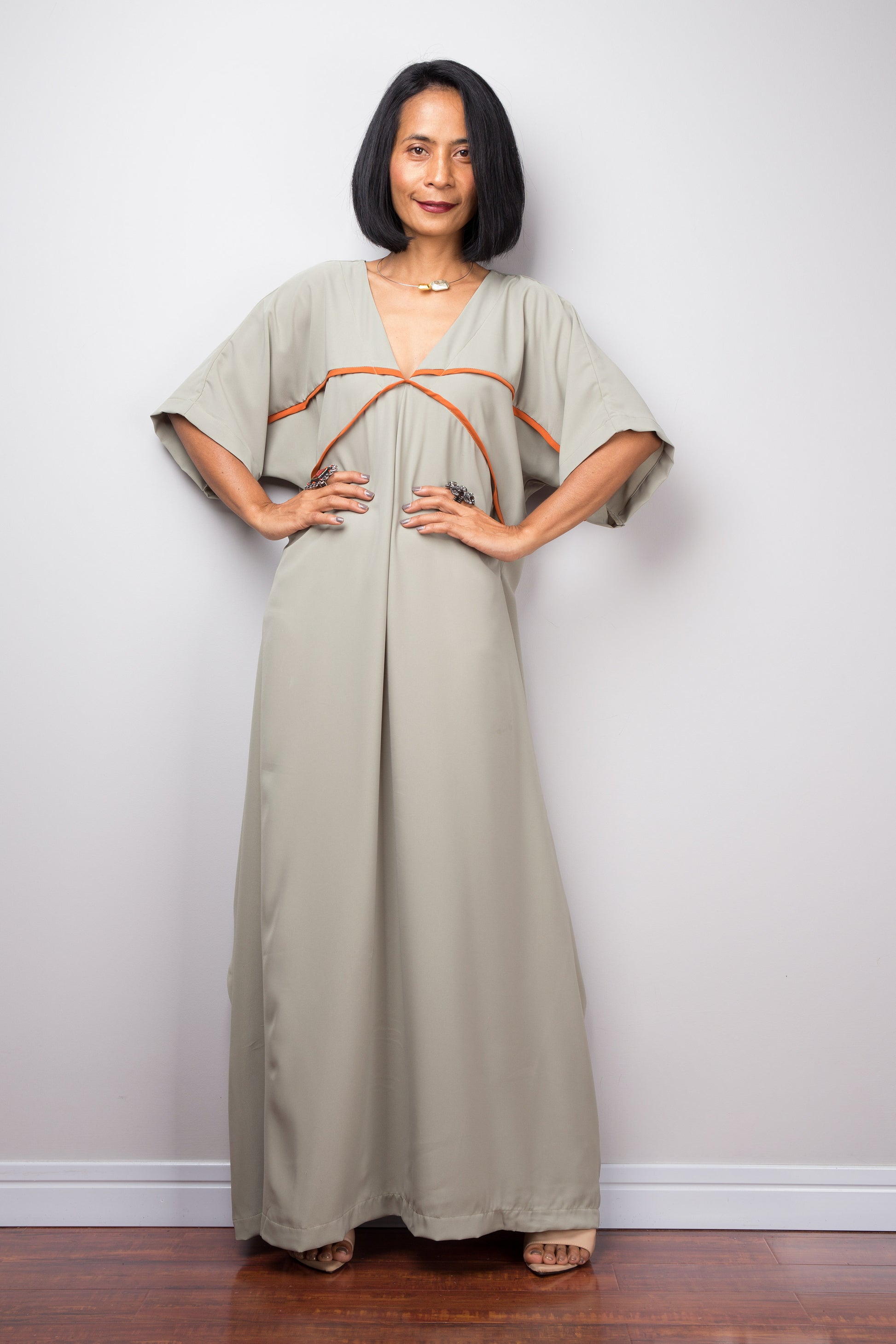 Woman in a sage green kaftan maxi dress by Nuichan, standing with hands on hips, showcasing wide sleeves and white trim, in a studio.

