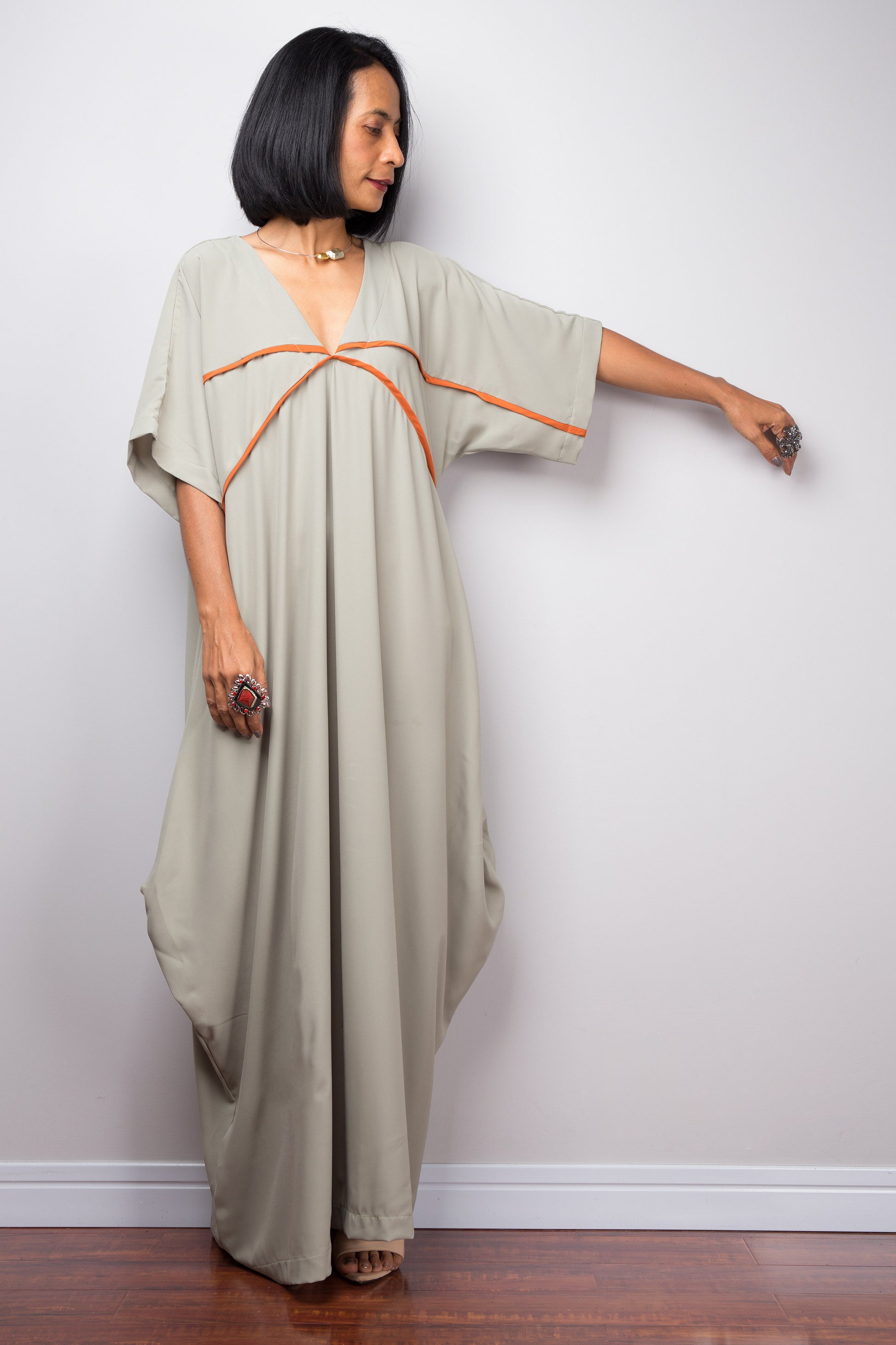 Nuichan’s sage kaftan maxi dress with white crisscross pattern, worn by a woman with dark hair, posed against a gray wall on a wooden floor, highlighting its short wide sleeves.

