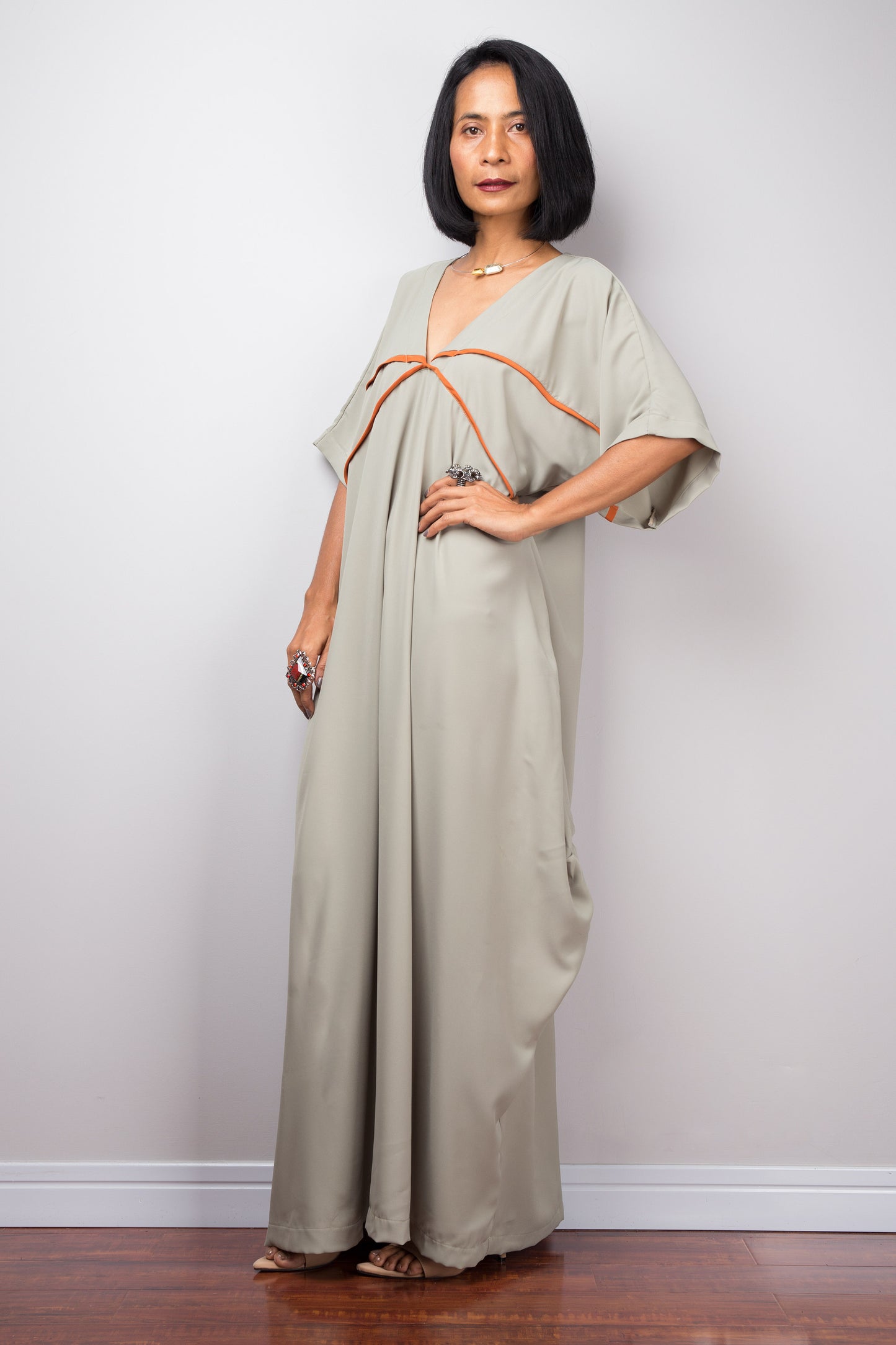 Woman in a sage kaftan maxi dress by Nuichan, standing with a hand on one hip, showcasing wide sleeves and white trim,  in a studio.