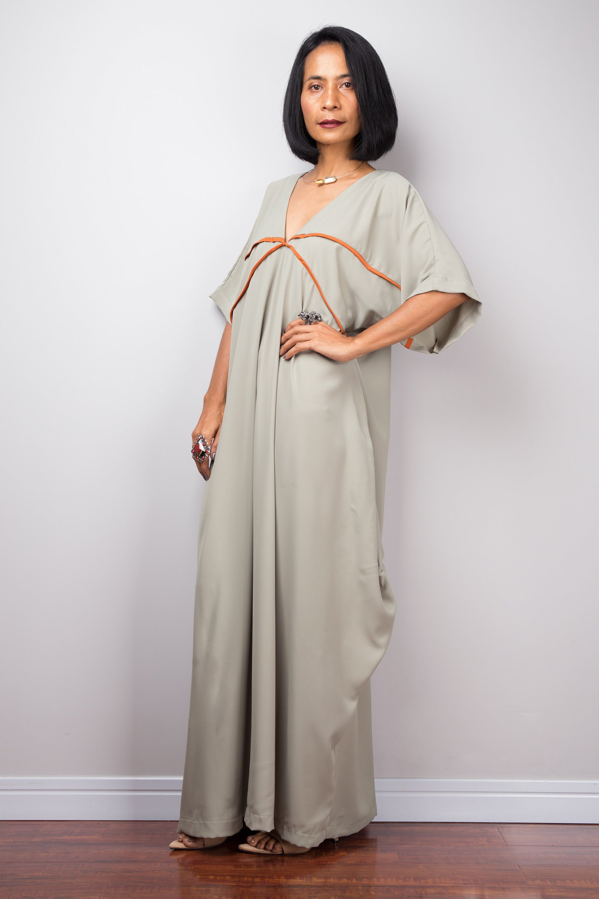 Woman in a sage kaftan maxi dress by Nuichan, standing with a hand on one hip, showcasing wide sleeves and white trim,  in a studio.