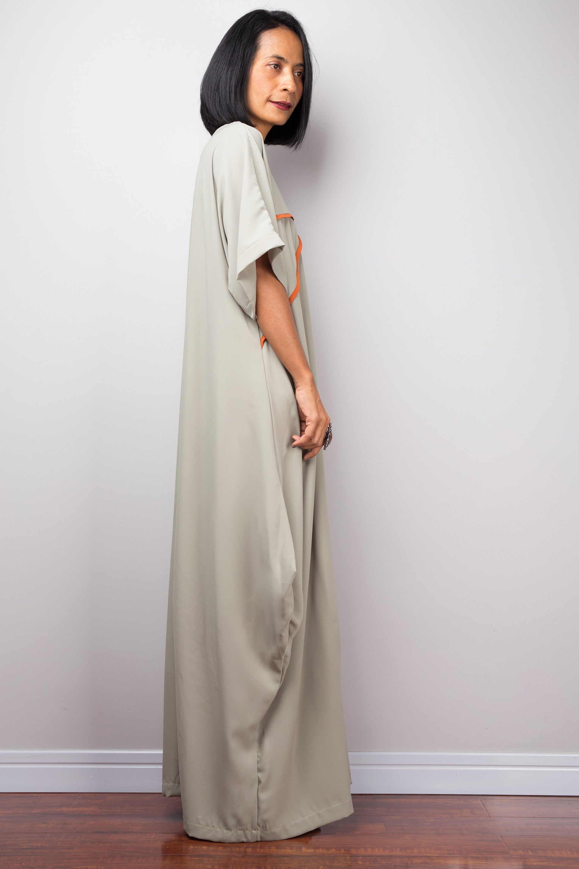 A floor-length sage gre kaftan dress from Nuichan, with white crisscross chest detail, worn by a woman standing sideways, set against a gray wall and wooden floor.